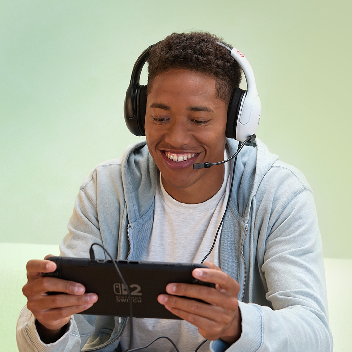 A smiling young man plays a Nintendo Switch, wearing a headset. He's in a grey hoodie, with a light green background.