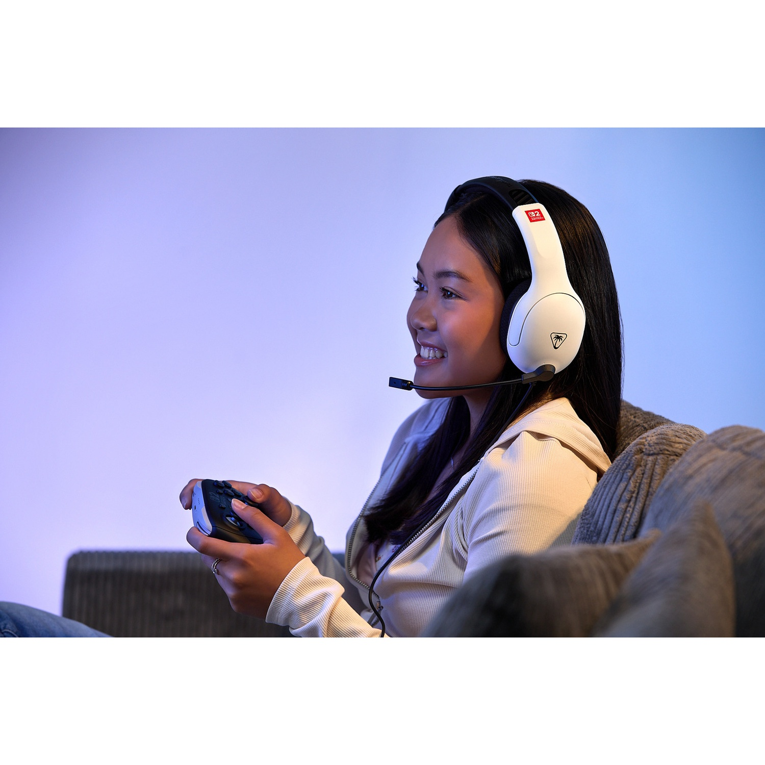 A young woman wearing a headset is smiling while holding a game controller. She's sitting on a couch with a blue and purple background.