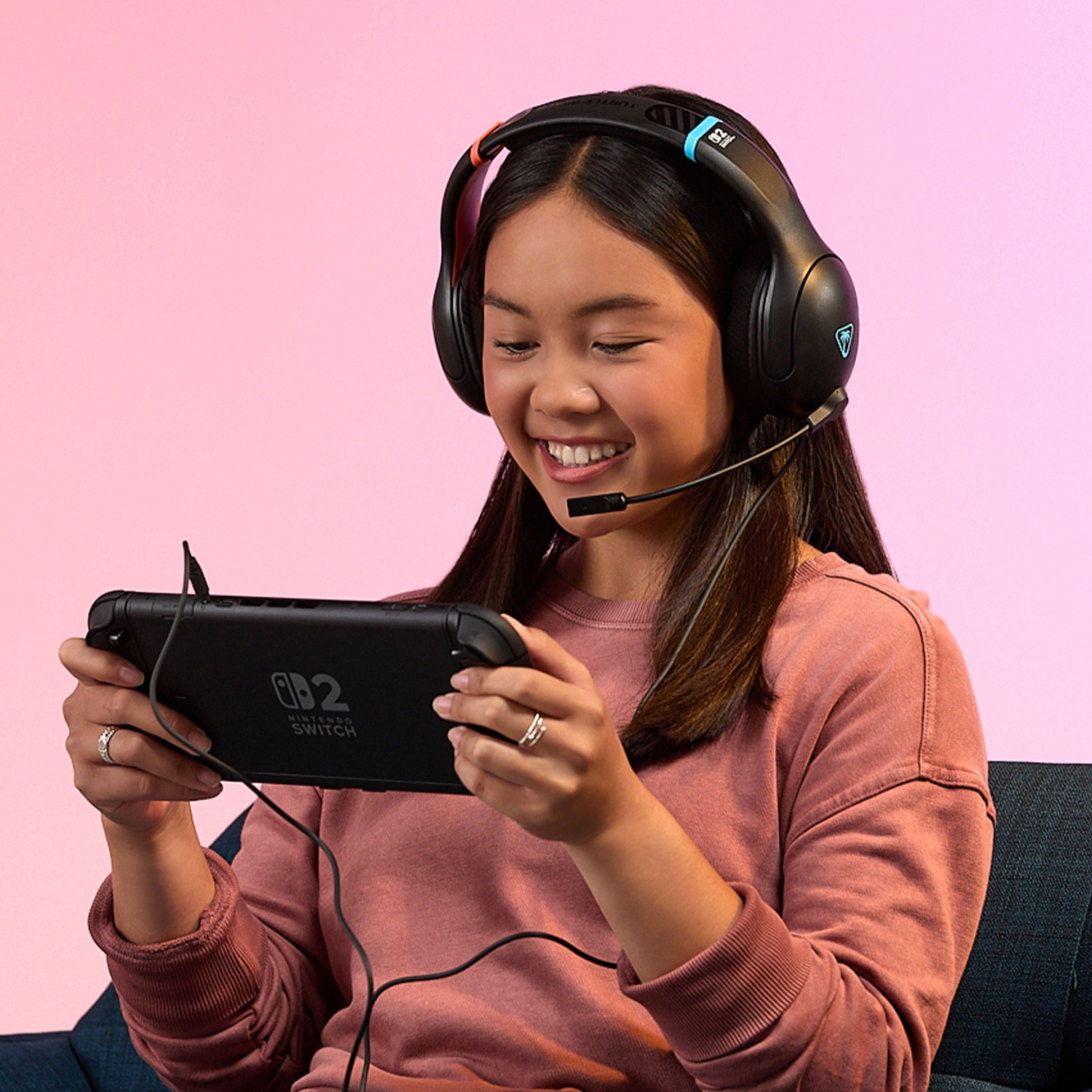A girl smiles, wearing a headset, holding a Nintendo Switch. Pink background. She is seated on a dark blue couch.