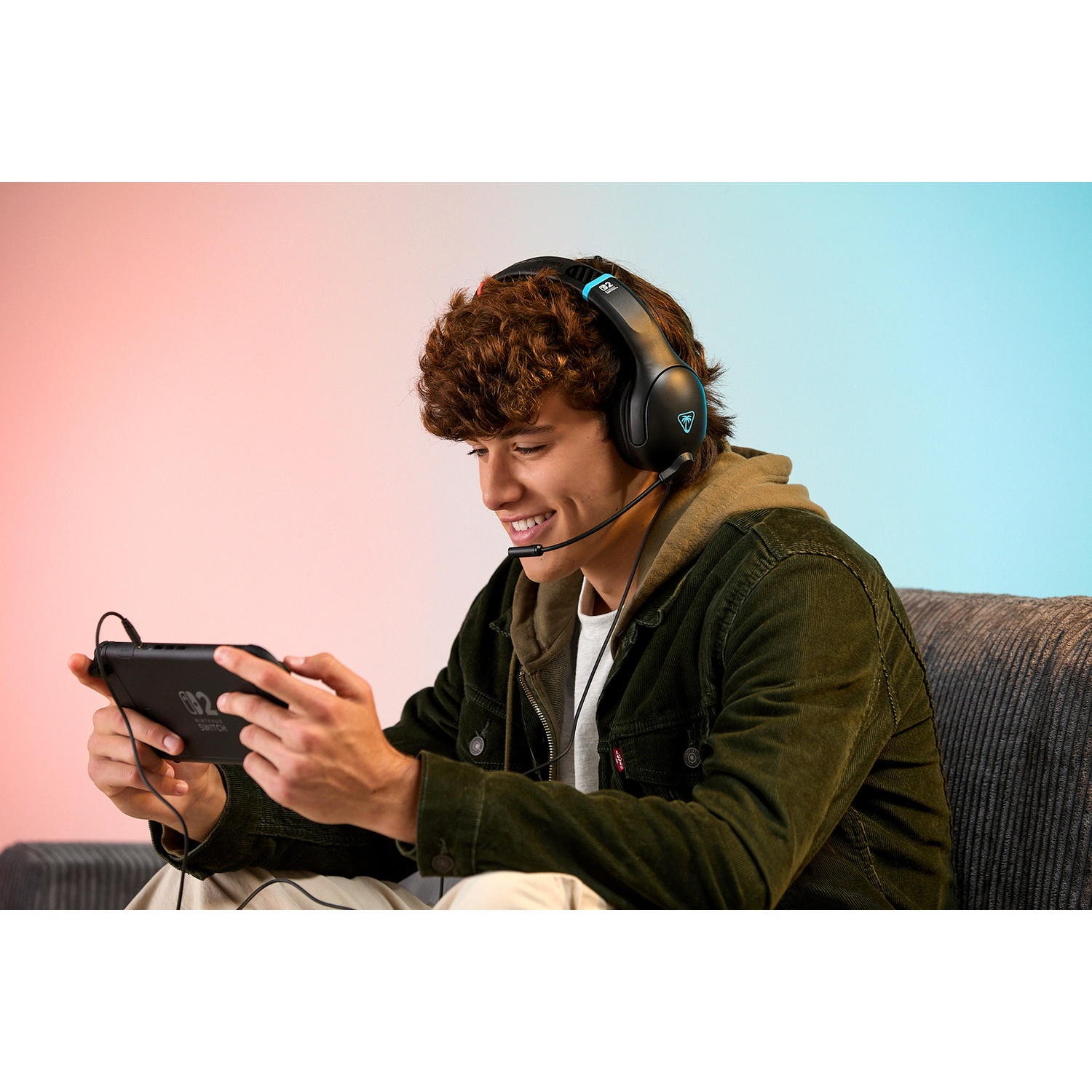 A young man wearing a headset plays a handheld game. He's smiling. The background is a mix of pink and blue.