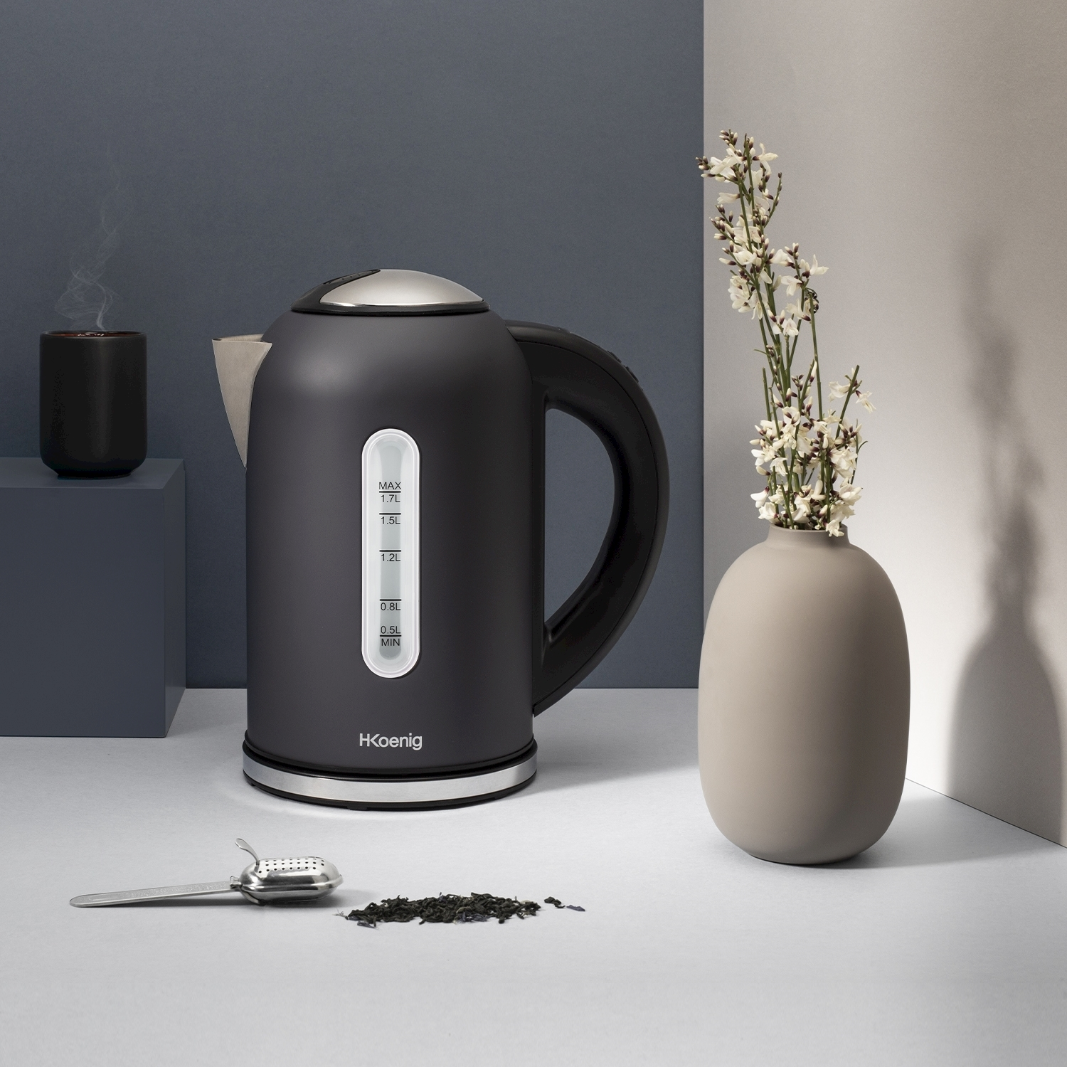 Un bollitore elettrico nero con una tazza, un vaso con fiori, foglie di tè e un infusore. Lo sfondo è un misto di grigio e bianco.