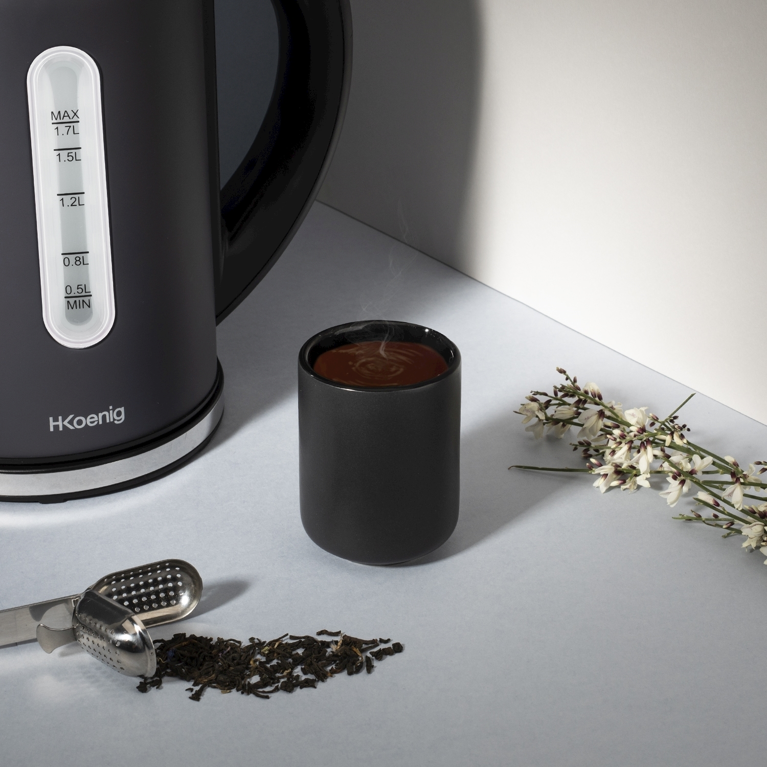 Un bollitore elettrico nero, una tazza di tè fumante, foglie di tè, un infusore e fiori disposti su una superficie chiara.