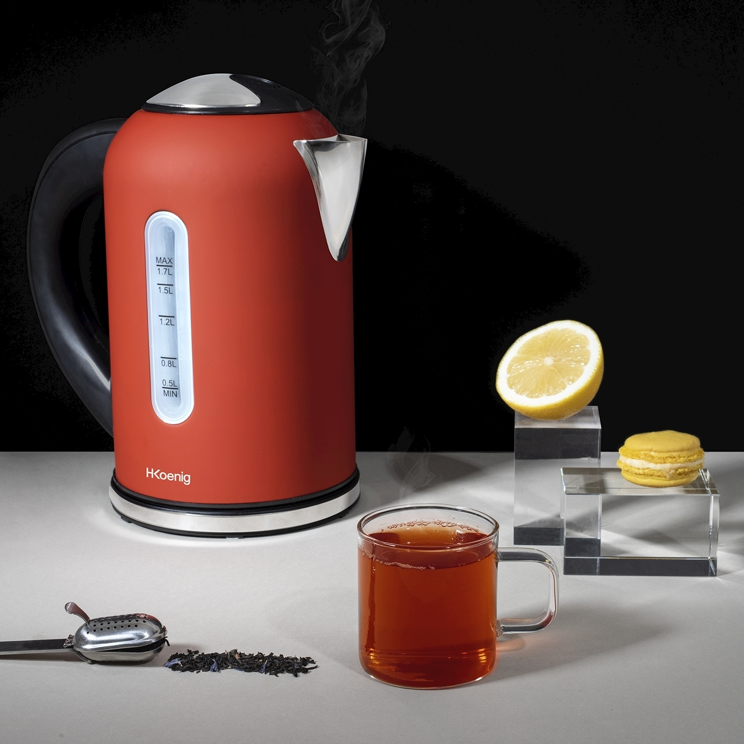 Bollitore rosso con vapore, tè in un bicchiere, limone e macaron su blocchi trasparenti. Sono presenti anche foglie di tè e un colino.