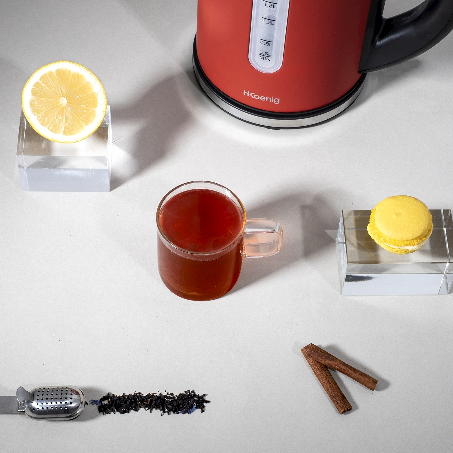 Natura morta con tè, limone, macaron, bastoncini di cannella, infusore e bollitore rosso su superficie bianca.