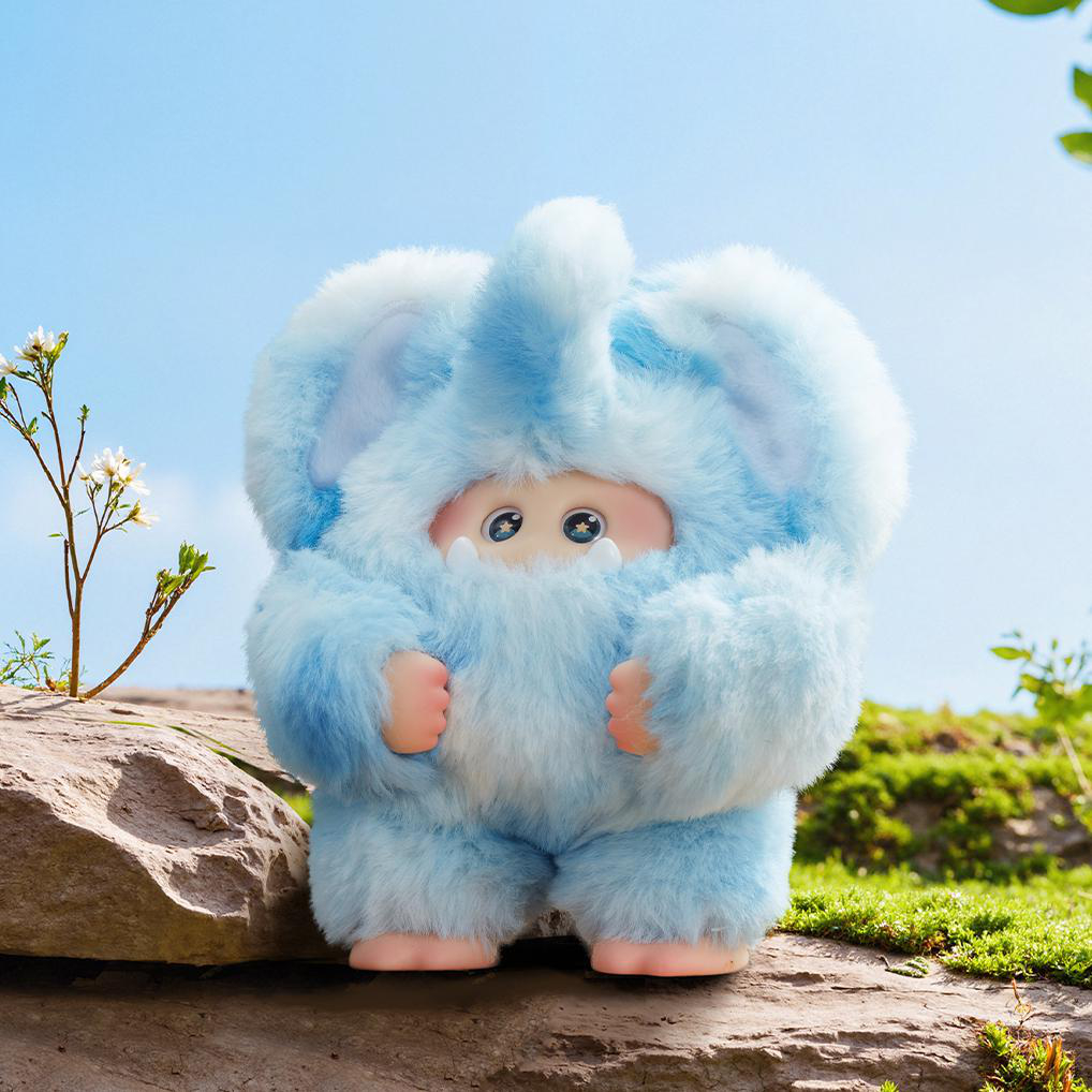 Eine plüschige, blaue, elefantenartige Figur steht auf einem Felsen, mit blauem Himmel im Hintergrund. Die Figur hat große Augen und kleine rosa Hände.
