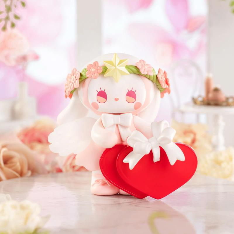 Eine Figur mit weißem Gesicht und Blumenkranz hält eine rot herzförmige Schachtel mit weißer Schleife. Weicher rosa Hintergrund.
