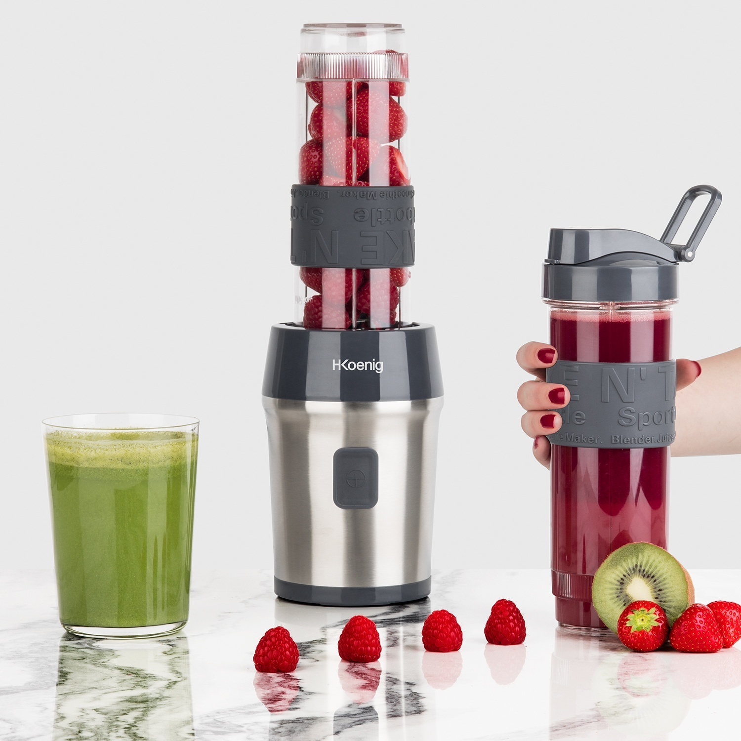 Un frullatore con lamponi, un frullato verde e un frullato con kiwi e fragole su una superficie di marmo. Una mano tiene un frullato.