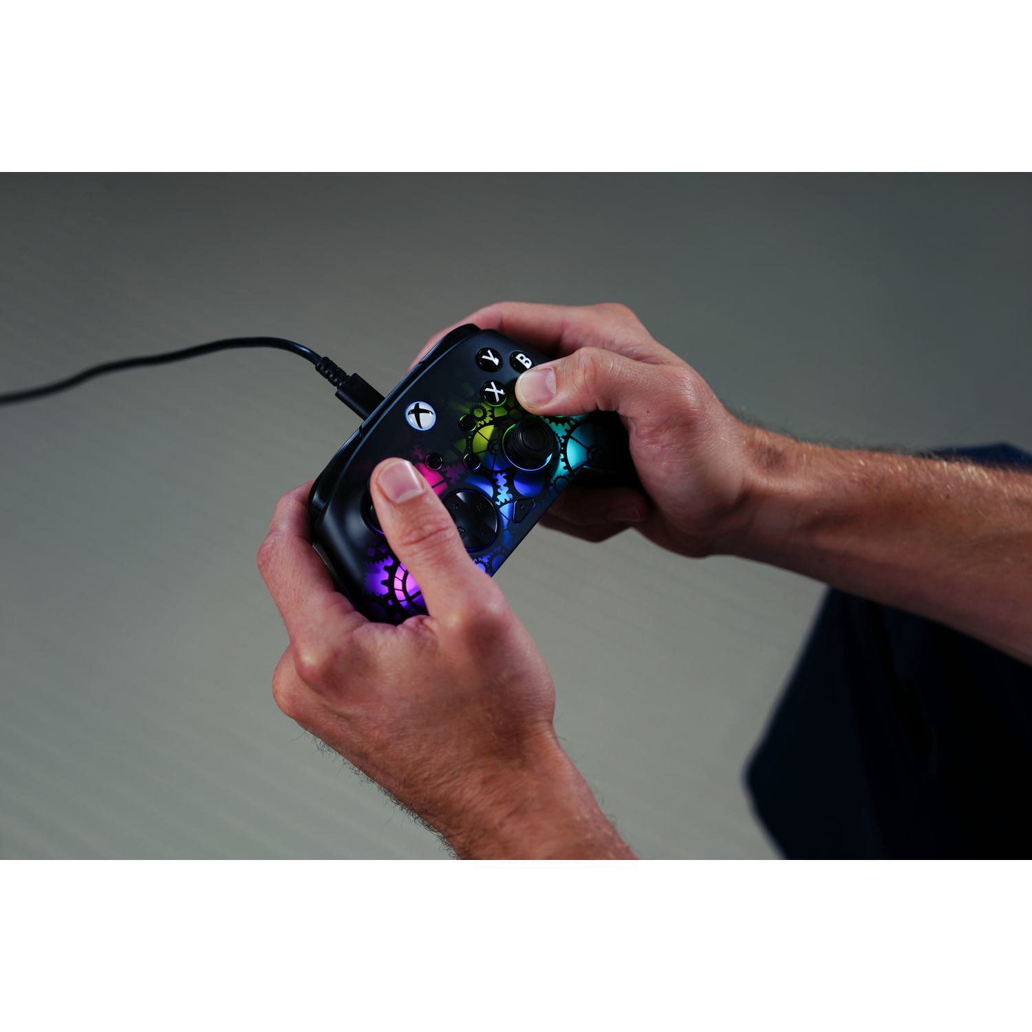 Hands holding a black Xbox controller with colorful lights and a USB cable. The background is a gradient gray.