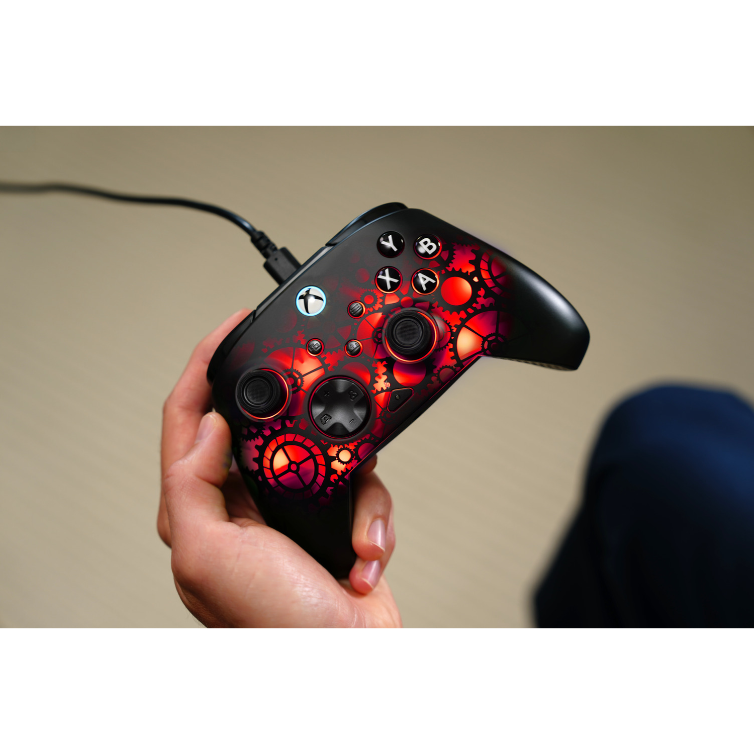 A hand holds a black Xbox controller with red gear designs. It is plugged in. The background is out of focus.