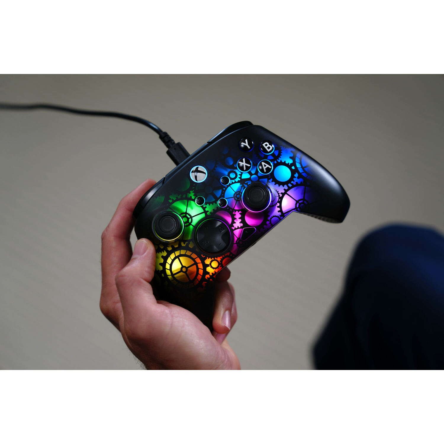 A hand holds a black Xbox controller with colorful gear designs. A cable is plugged in. The background is blurred.