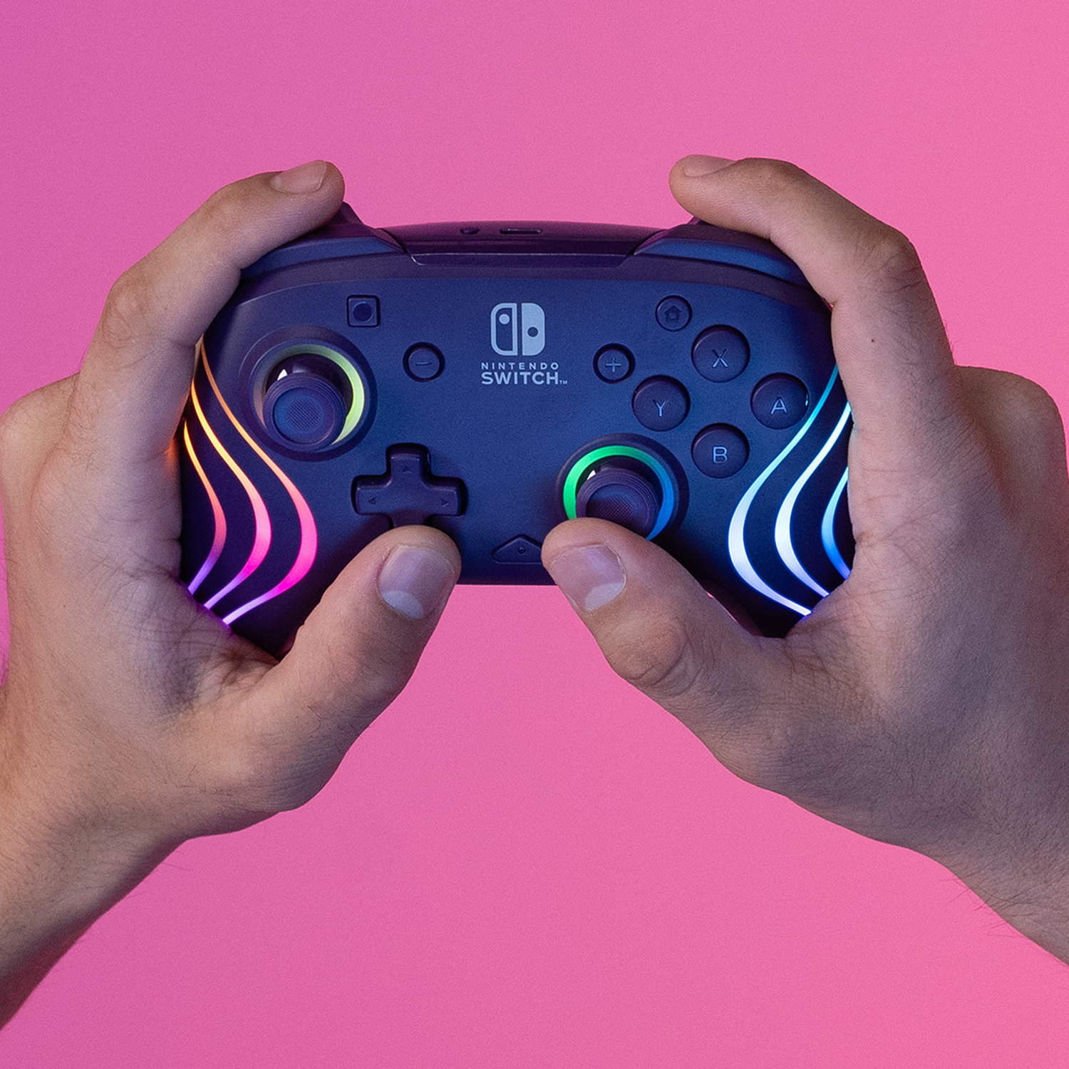 Handen houden een Nintendo Switch-controller vast. Deze is donkerblauw met gekleurde lampjes en knoppen, op een roze achtergrond.