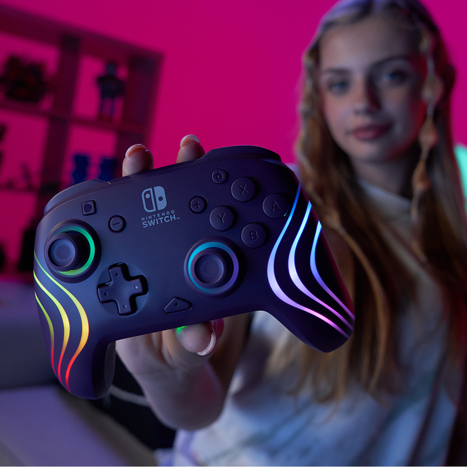 Een persoon houdt een Nintendo Switch-controller vast met kleurrijke lichten. De achtergrond toont een kamer en een andere persoon, enigszins onscherp.