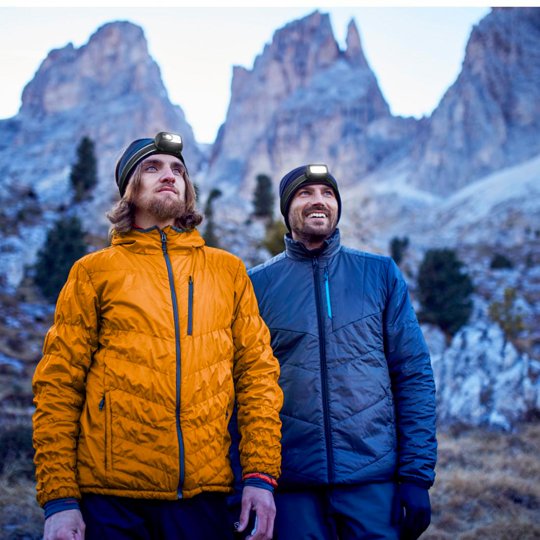 Zwei Männer in Jacken und Stirnlampen blicken auf Berggipfel. Einer trägt Orange, der andere Blau, beide mit Mützen.