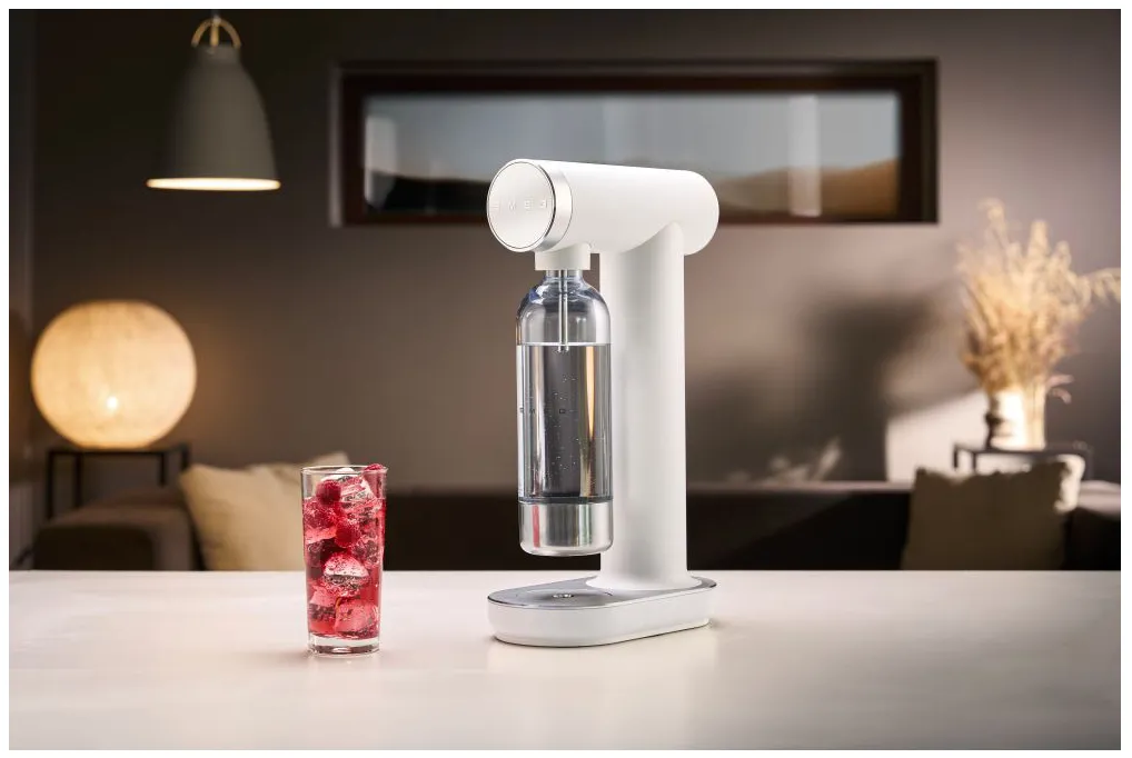 Un produttore di soda bianco con un bicchiere di bevanda rossa. Una lampada e una pianta decorativa sono sullo sfondo.