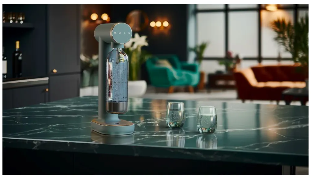 Un appareil à soda, deux verres d'eau avec des bulles sur un comptoir en marbre. L'arrière-plan présente un salon avec un canapé.