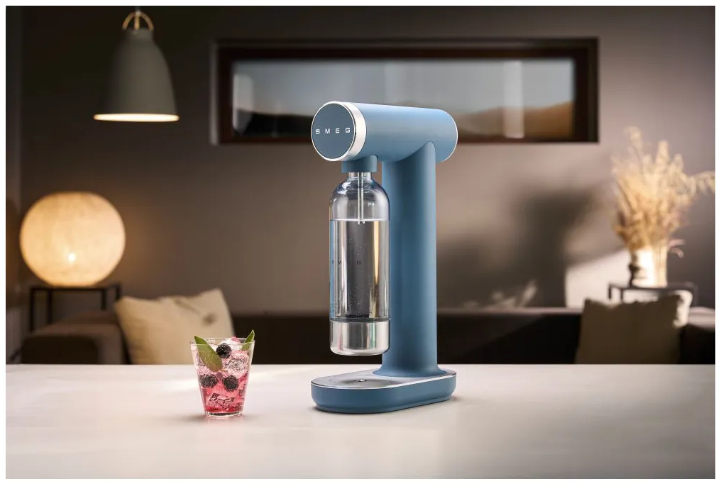 Blauer SMEG-Sodabereiter mit Glasflasche, Getränk und Raumumgebung. Dekorative Lampe und Fenster im Hintergrund.