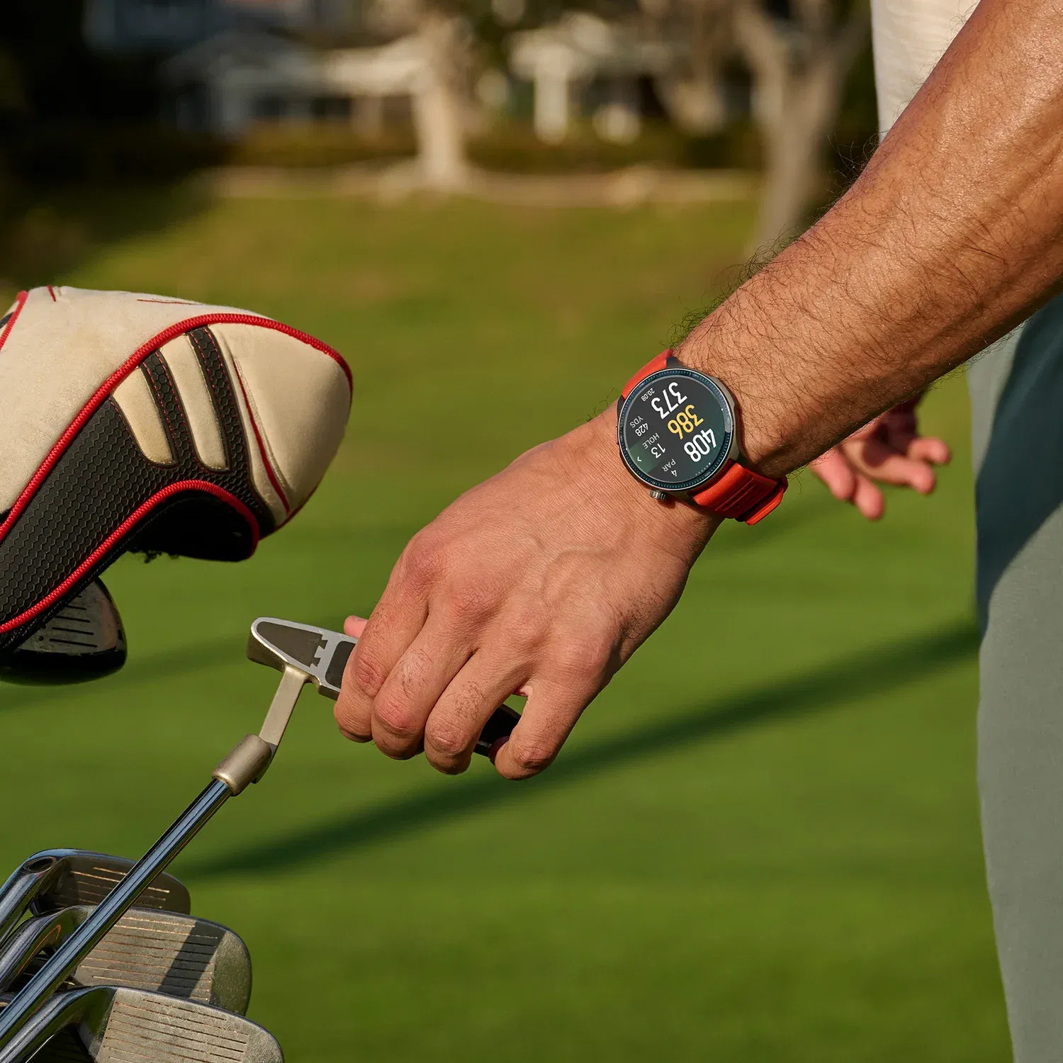 Golfista trzymający putter, noszący smartwatch. Widoczne kije golfowe i torba golfowa. Zielona trawa wypełnia tło.
