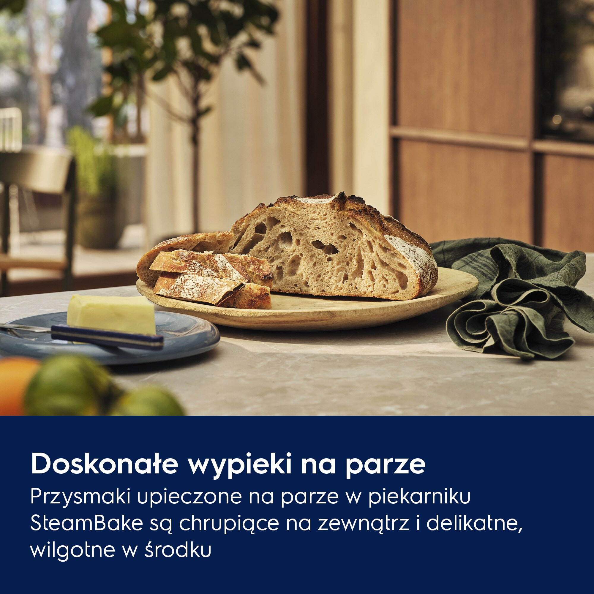 Kawałki chleba na drewnianym talerzu, masło, nóż i zielona serwetka na stole. W tle drzewo i okno.