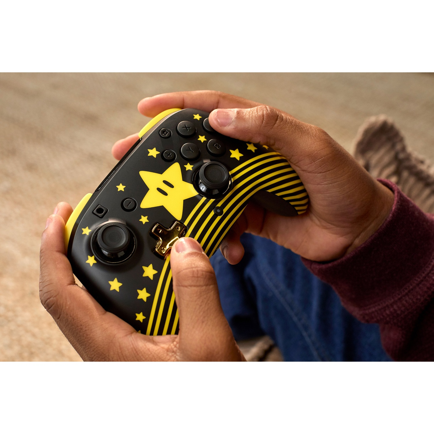 Mains tenant une manette de jeu noire et jaune sur le thème de Mario. Des étoiles et des rayures la décorent. La personne porte un jean bleu et une manche marron.