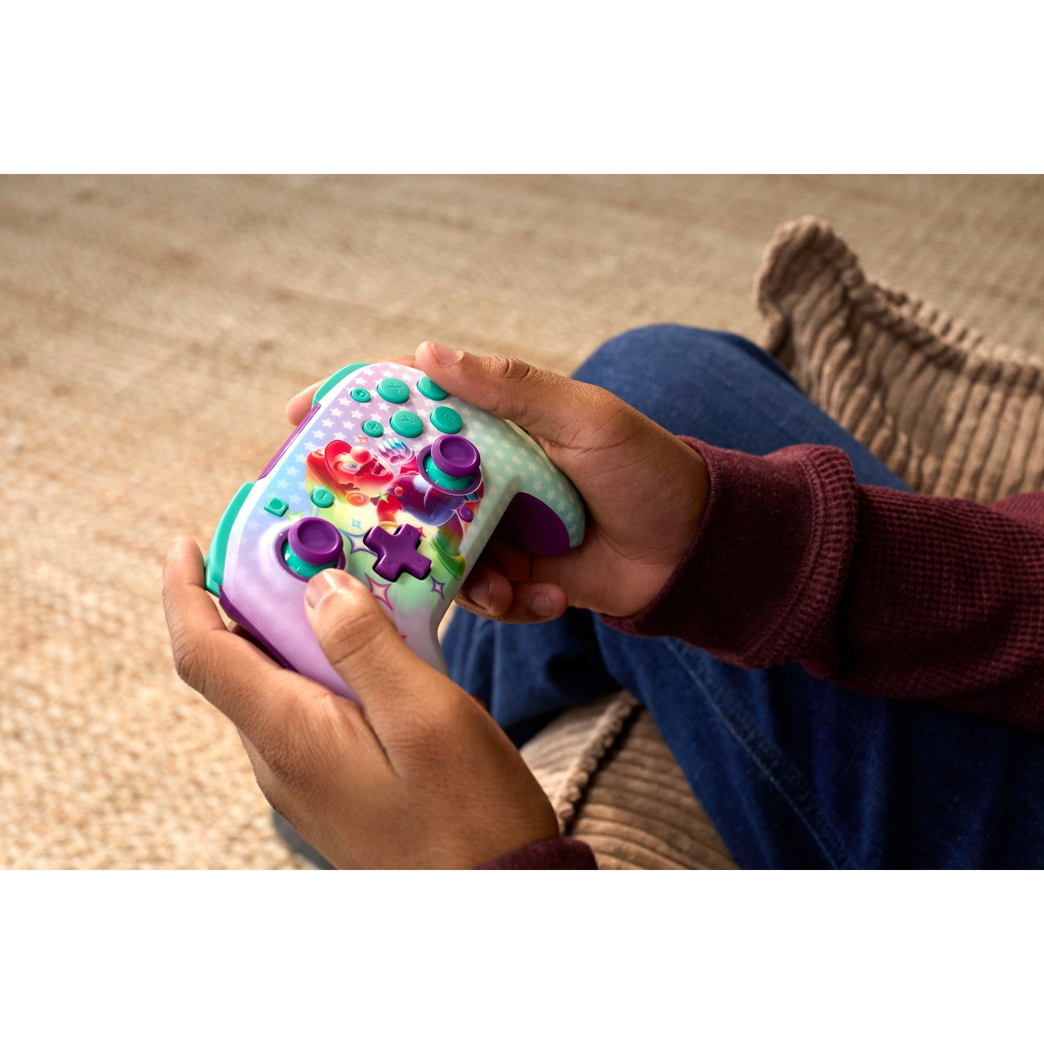 Une personne tient une manette de jeu colorée avec des dessins animés. Elle est assise, portant un pull bordeaux et un jean.