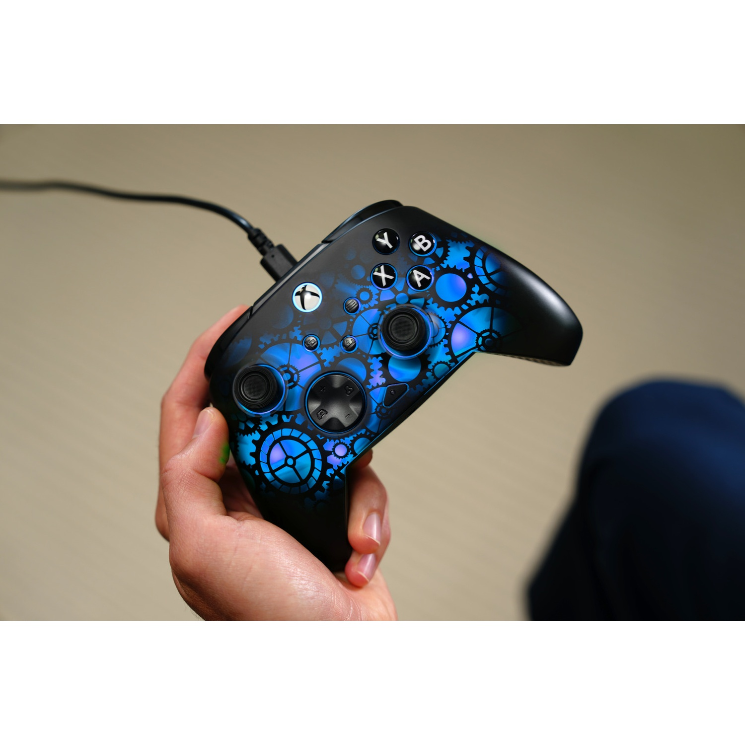 A person holding a black Xbox controller with a blue gear design. A cable connects to the controller. A person's leg is in the background.