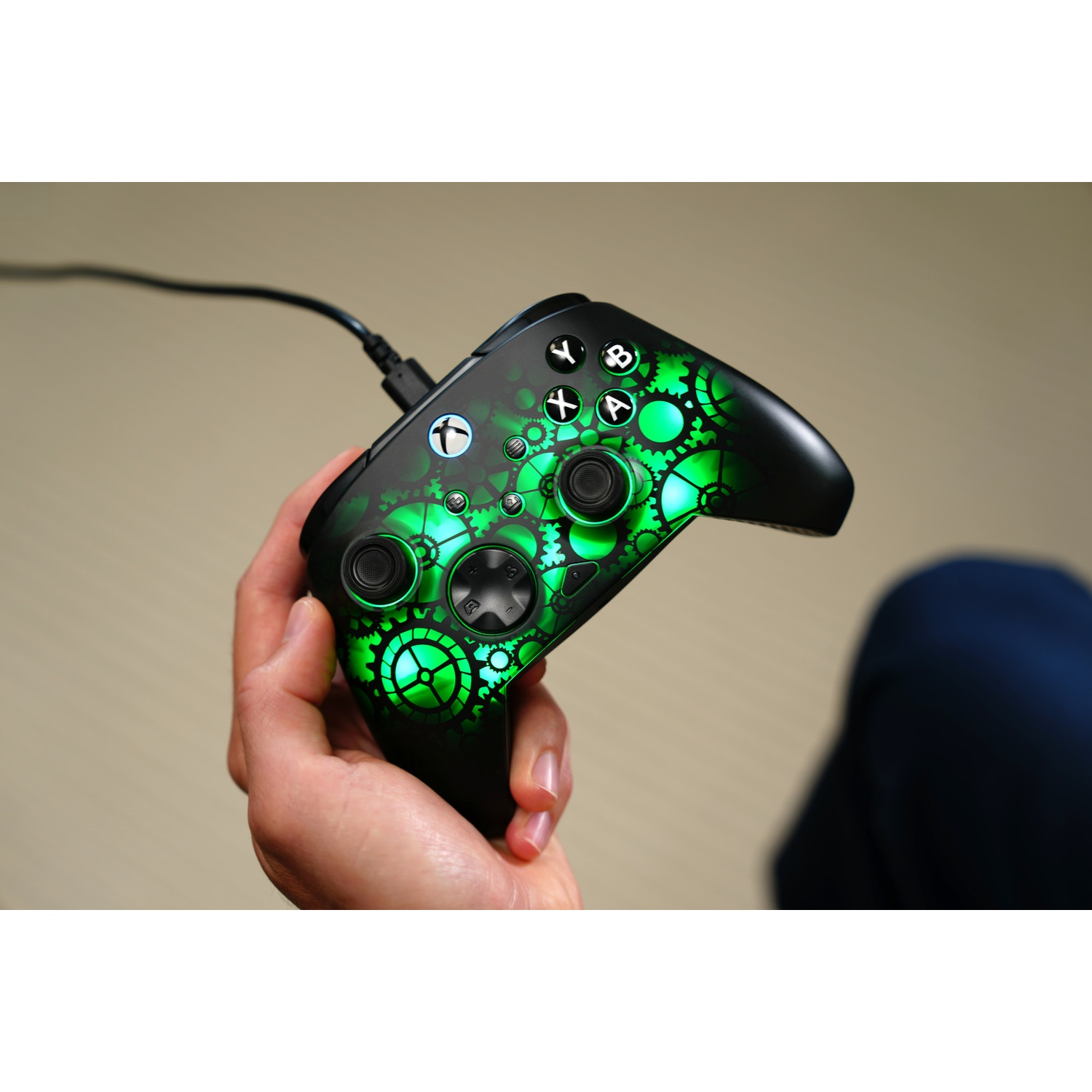 A person holding a black Xbox controller with green gear design. Controller is plugged in. Background is a light beige.