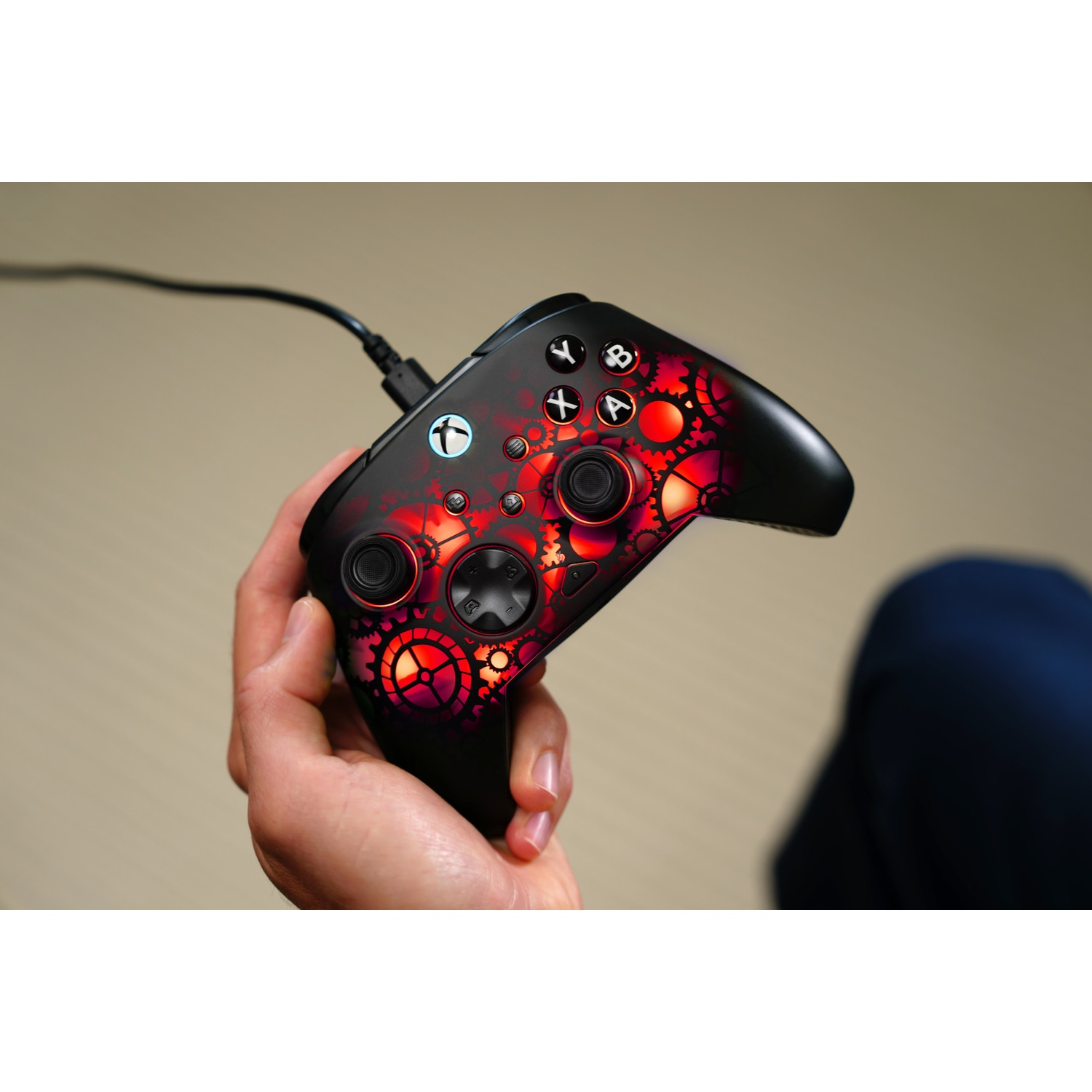 A person holds a black and red Xbox controller with a gear design. A cable is plugged in. The background is blurred.