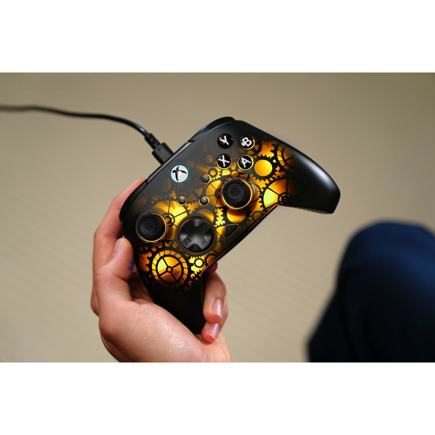 A hand holds a black Xbox controller with a steampunk gear design. The controller is connected by a black cable.