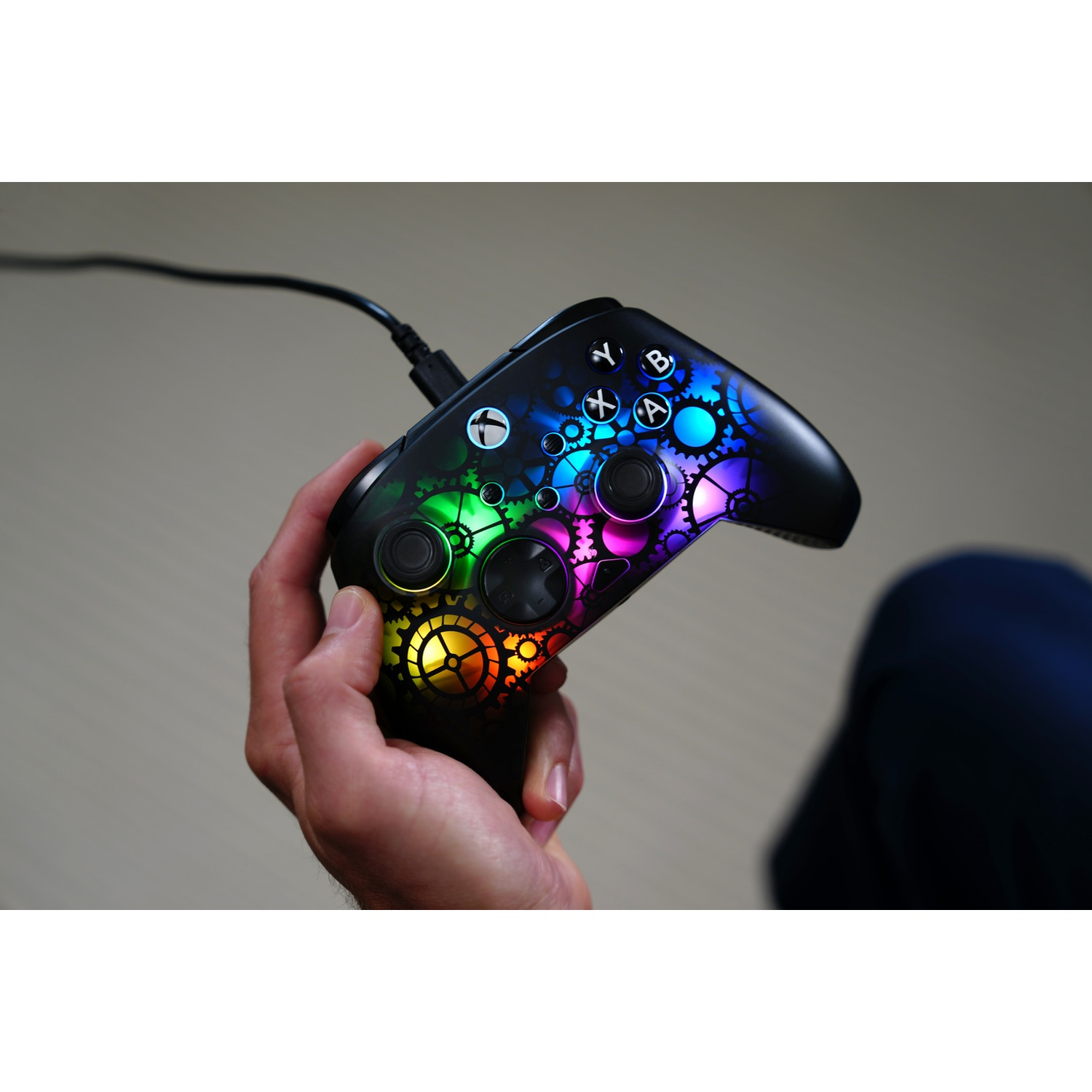 A hand holds a black Xbox controller with colorful gear designs. A cable is plugged in. The background is a blurred beige and blue.