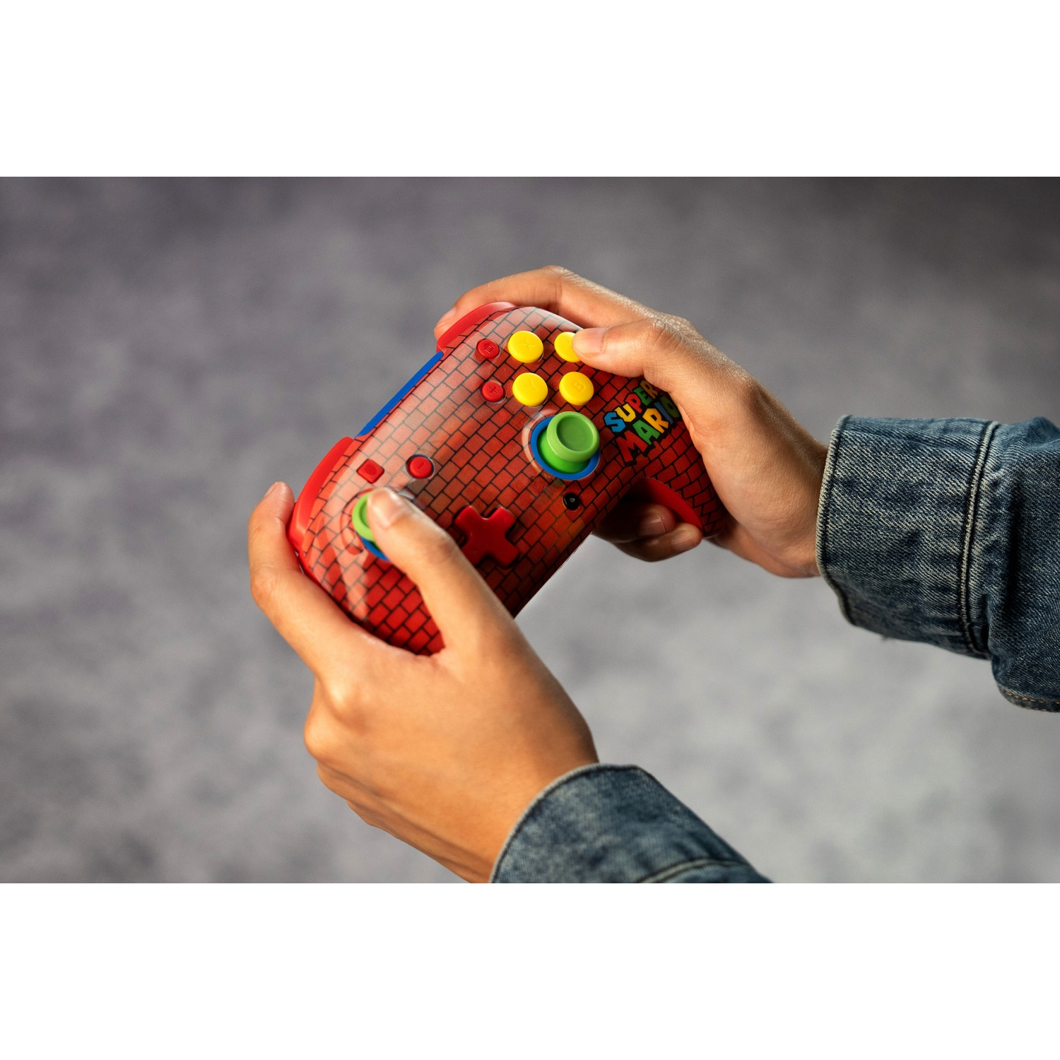 Mains tenant une manette Super Mario à motif de briques rouges. Des boutons jaunes, verts et rouges sont visibles. Des manches de veste en jean sont également visibles.