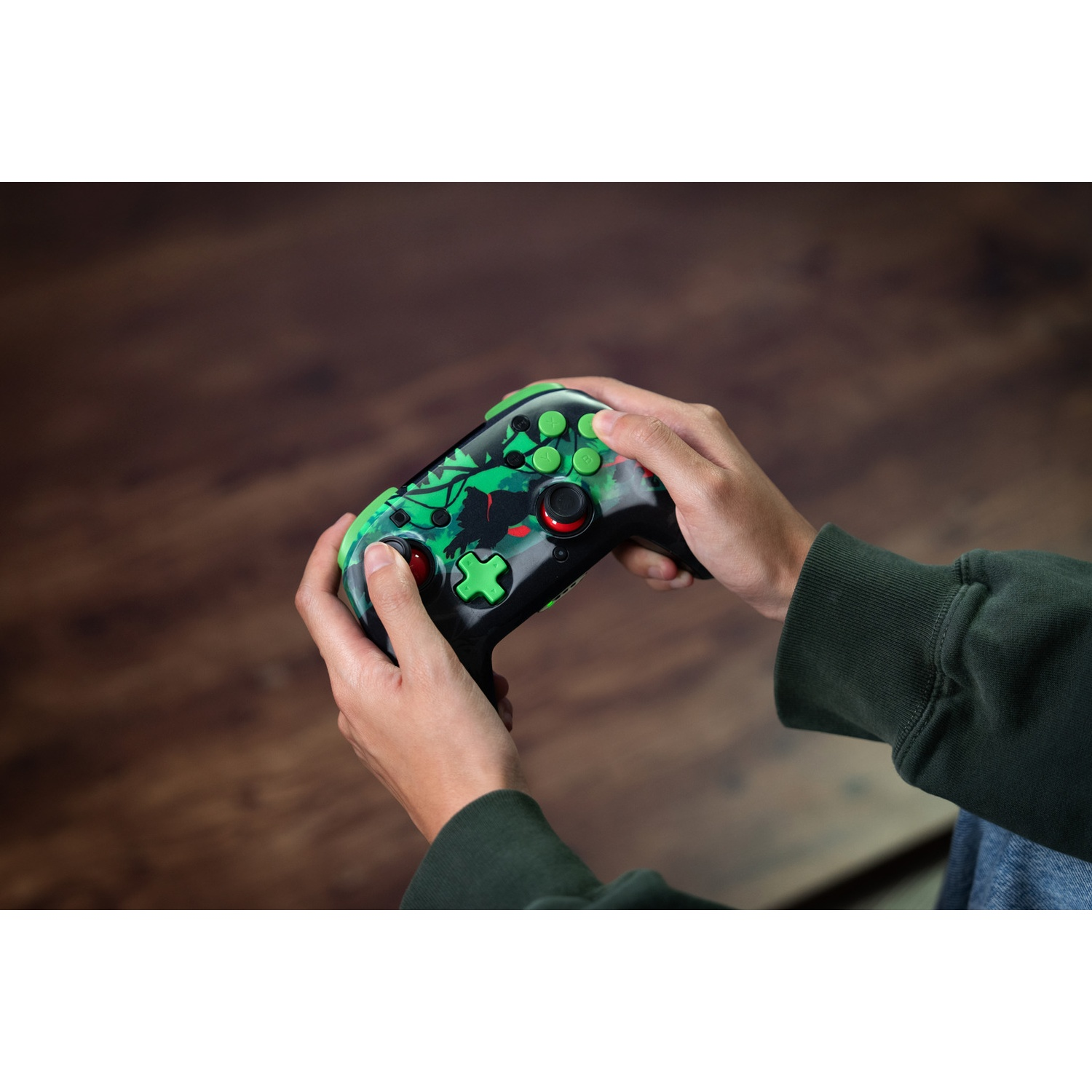 Mains tenant une manette de jeu noire et verte, avec des boutons verts et des touches rouges, sur fond de bois.