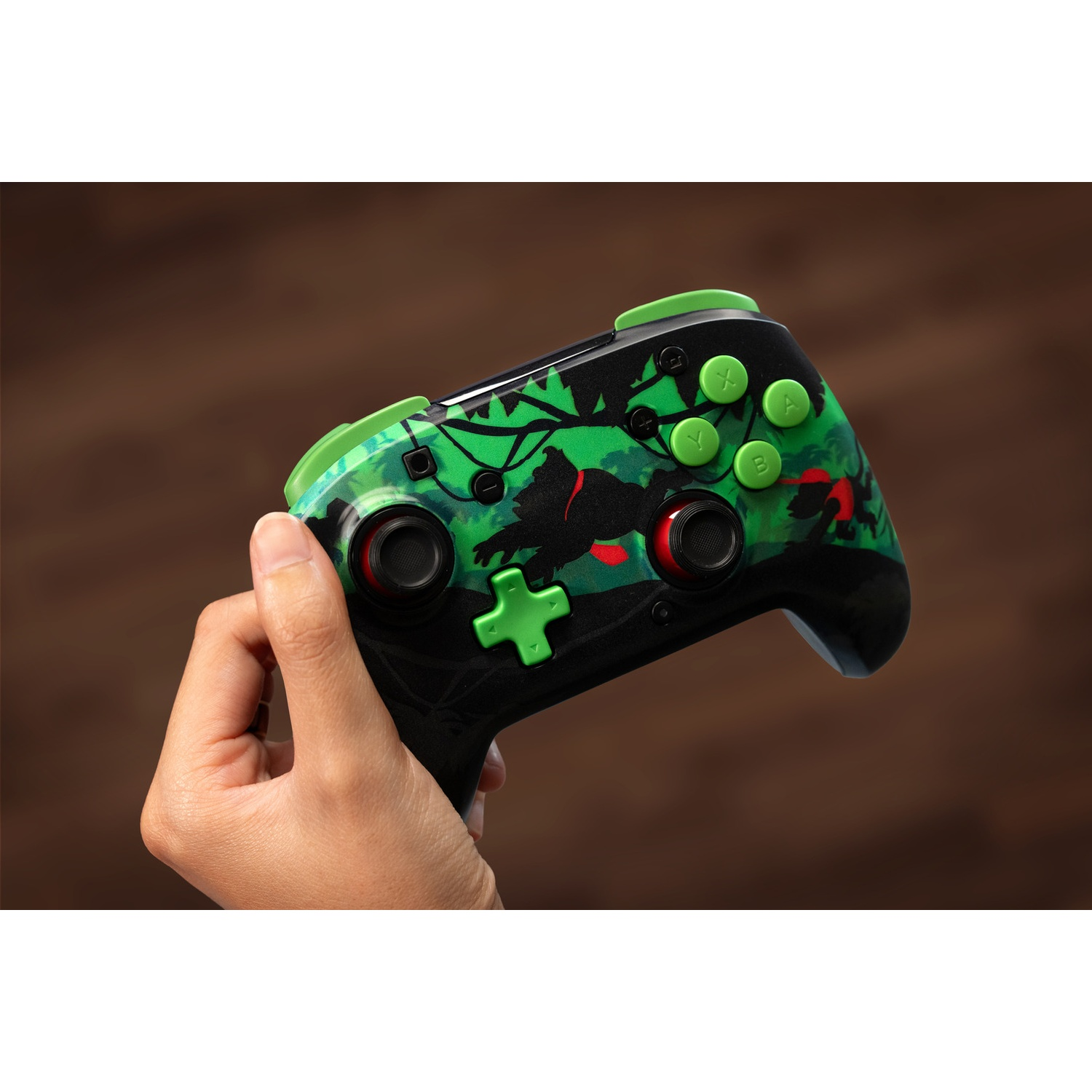 Une main tenant une manette de jeu vidéo noire et verte. L'arrière-plan est une surface en bois marron floue.