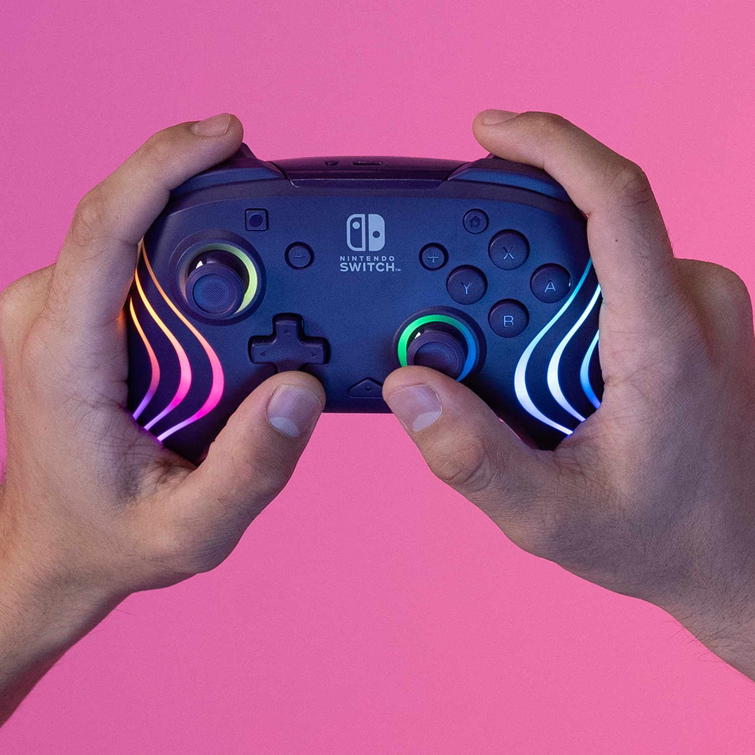 Hands holding a Nintendo Switch controller. It has colorful lights and buttons. Pink background.