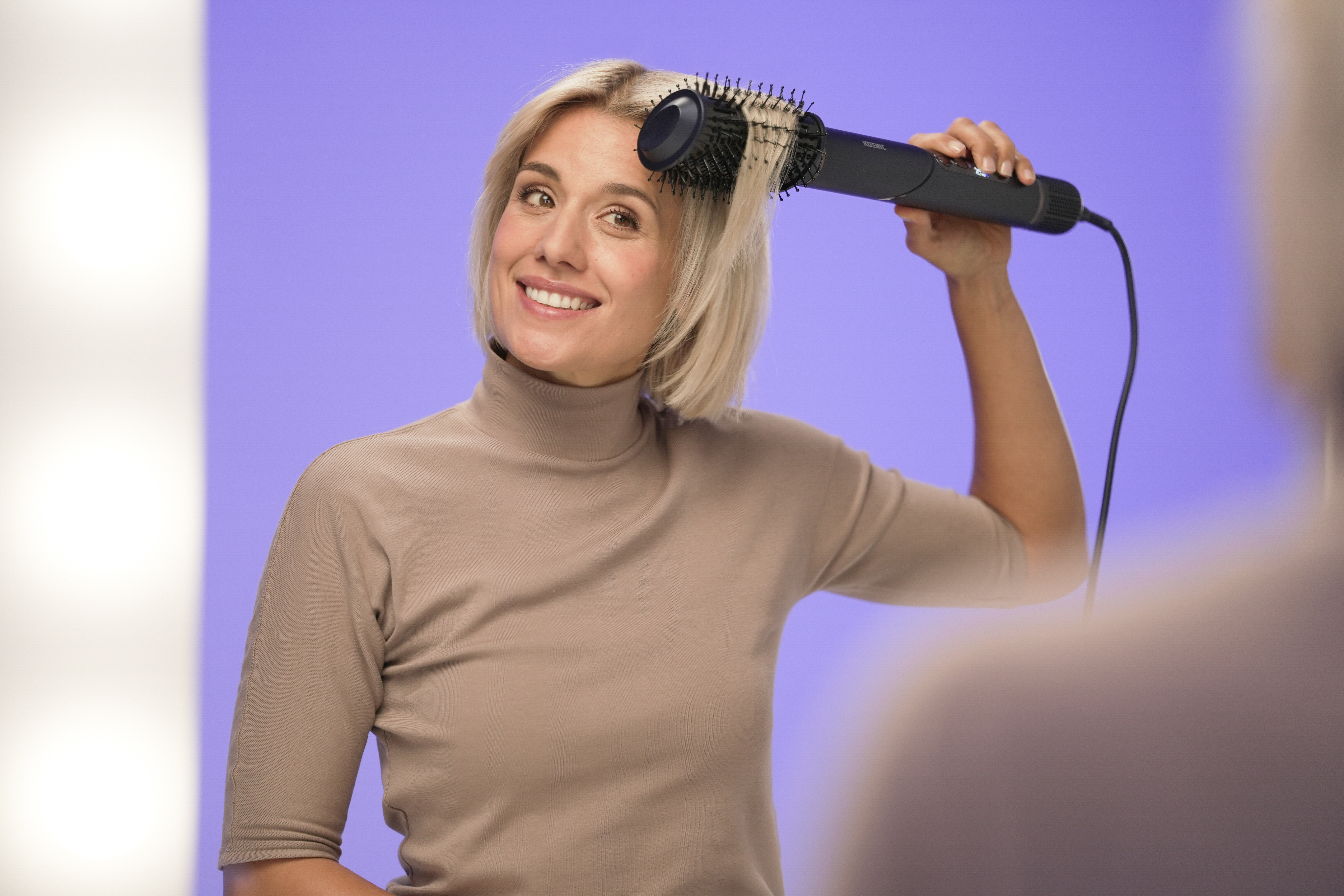 Eine Frau frisiert ihre Haare mit einer schwarzen Haartrocknerbürste. Sie lächelt. Der Hintergrund ist lila.
