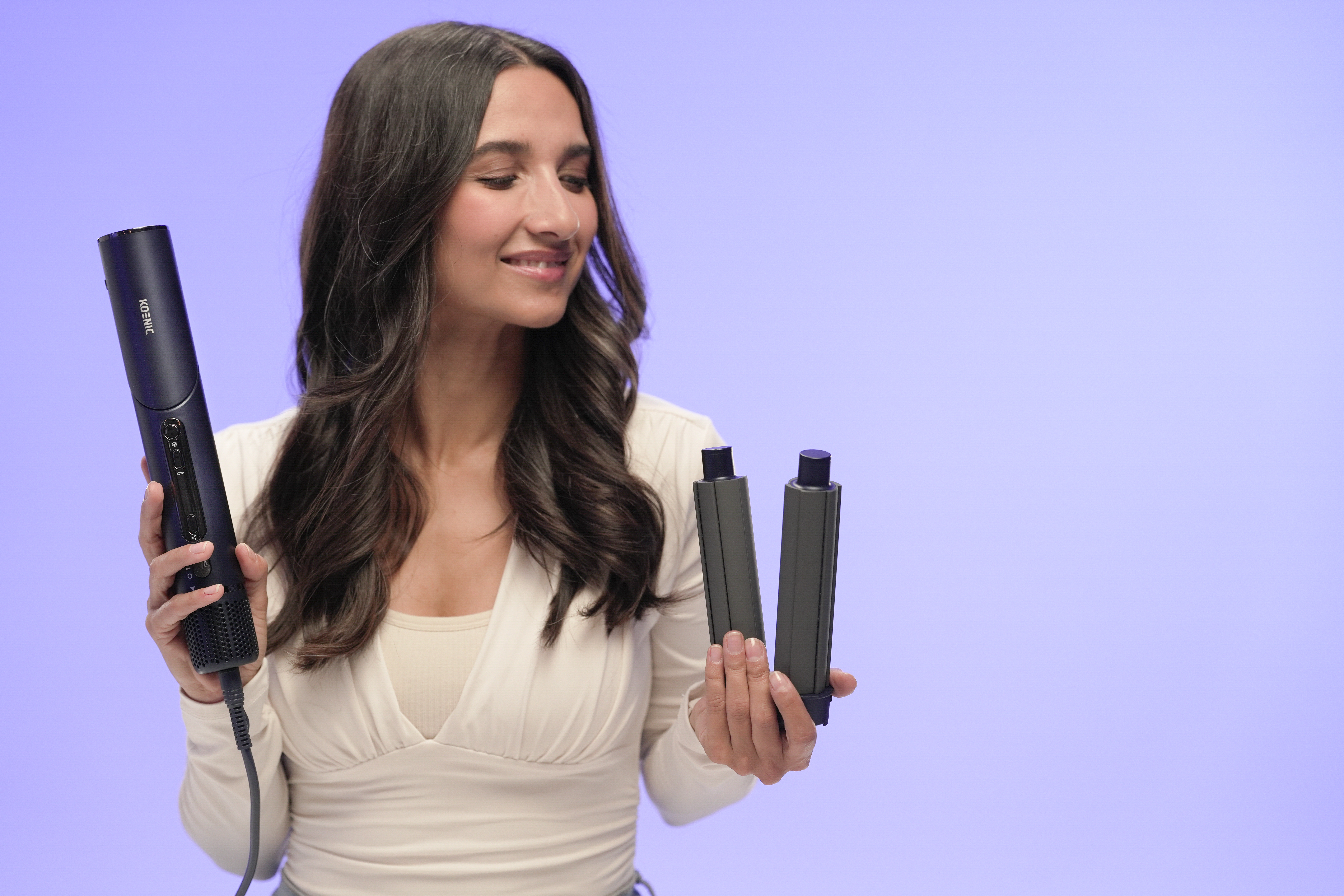 Frau hält Haarstyling-Tools und lächelt. Sie hält einen Haartrockner und zwei Aufsätze vor einem lilafarbenen Hintergrund.