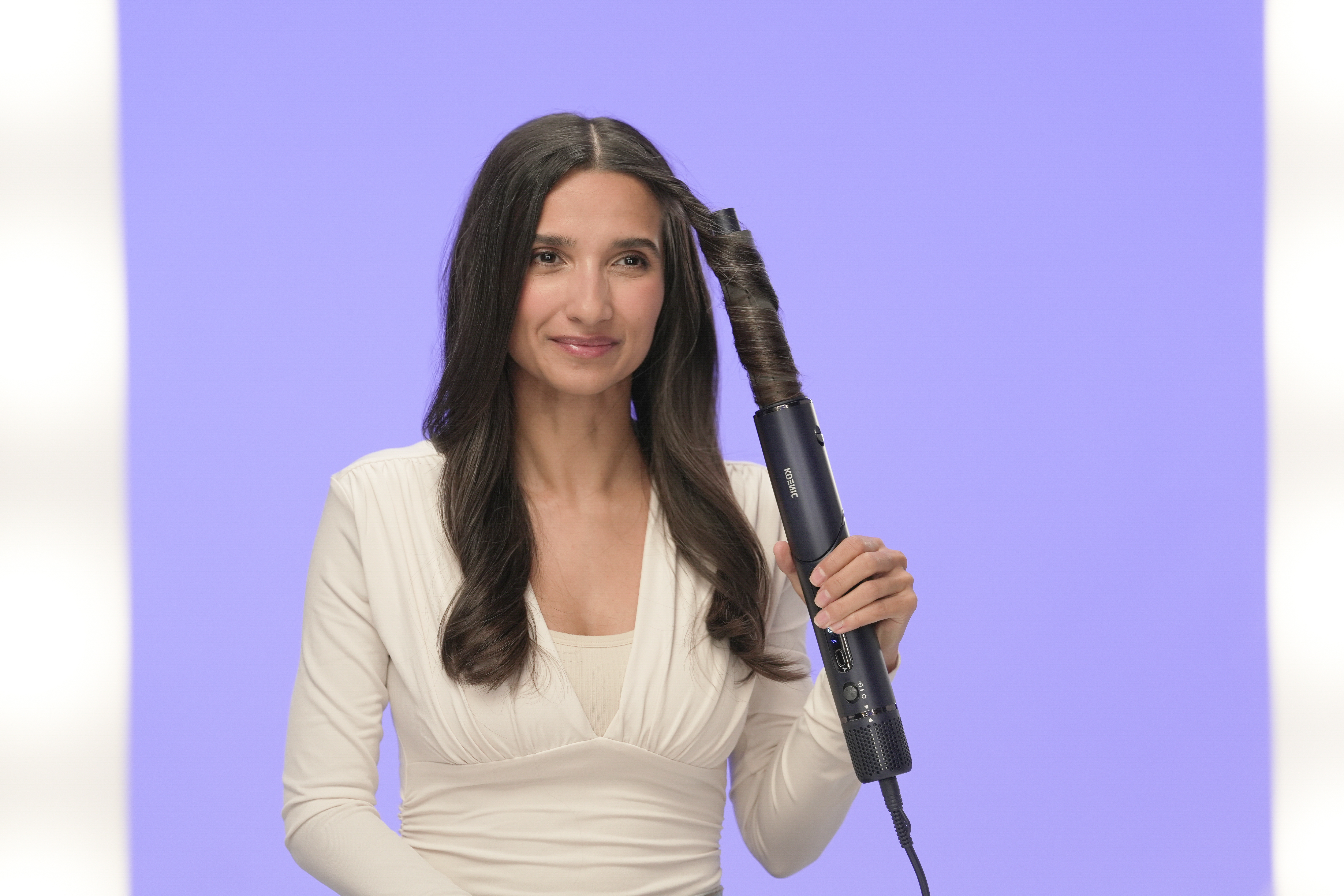 Eine Frau lockt ihr Haar mit einem dunkelblauen Lockenstab. Sie trägt ein weißes Oberteil. Der Hintergrund ist lila und weiß.