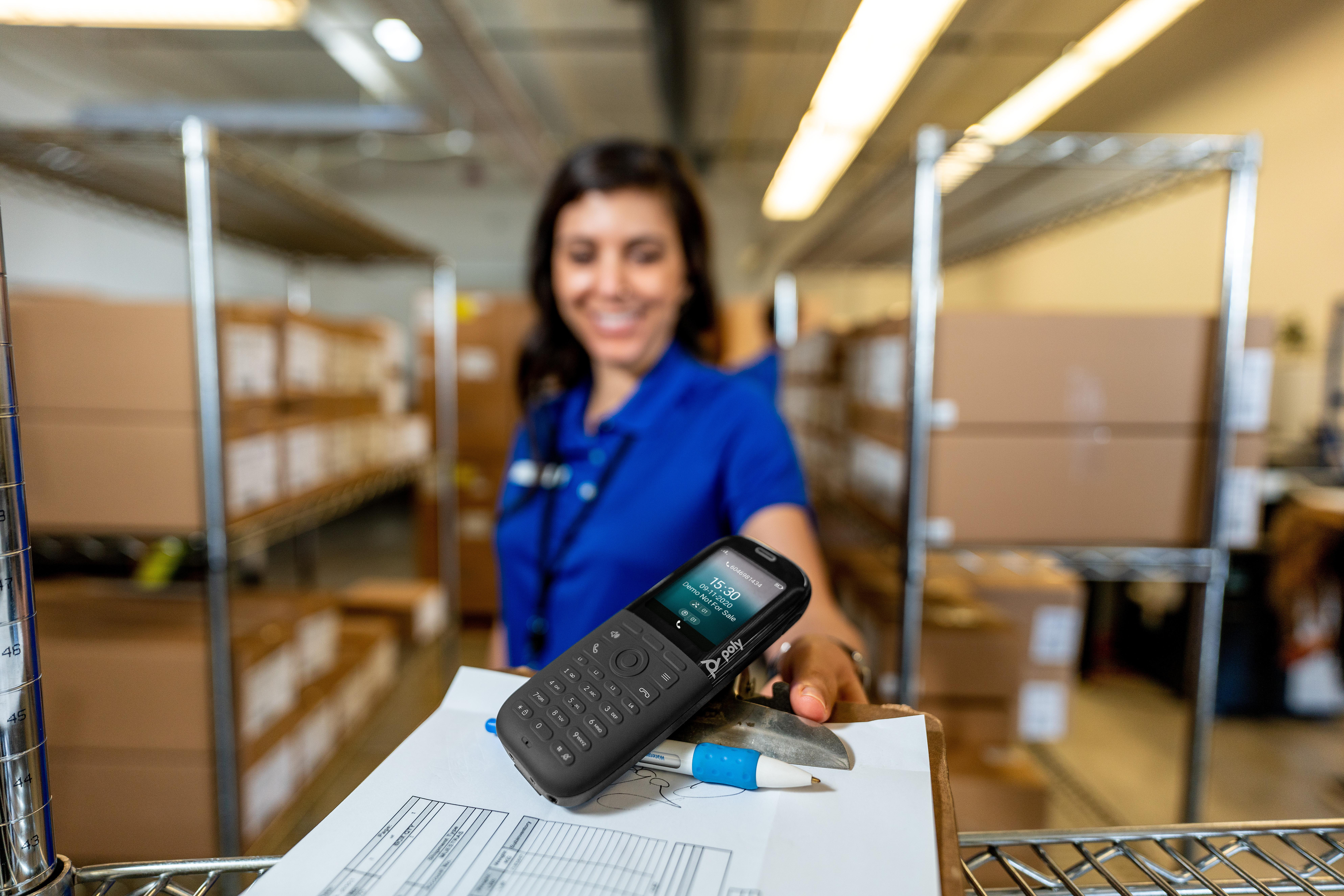 Eine Frau in Blau bietet ein Telefon mit einem Bildschirm an, der Informationen anzeigt. Stift und Papier liegen davor. Regale sind im Hintergrund.