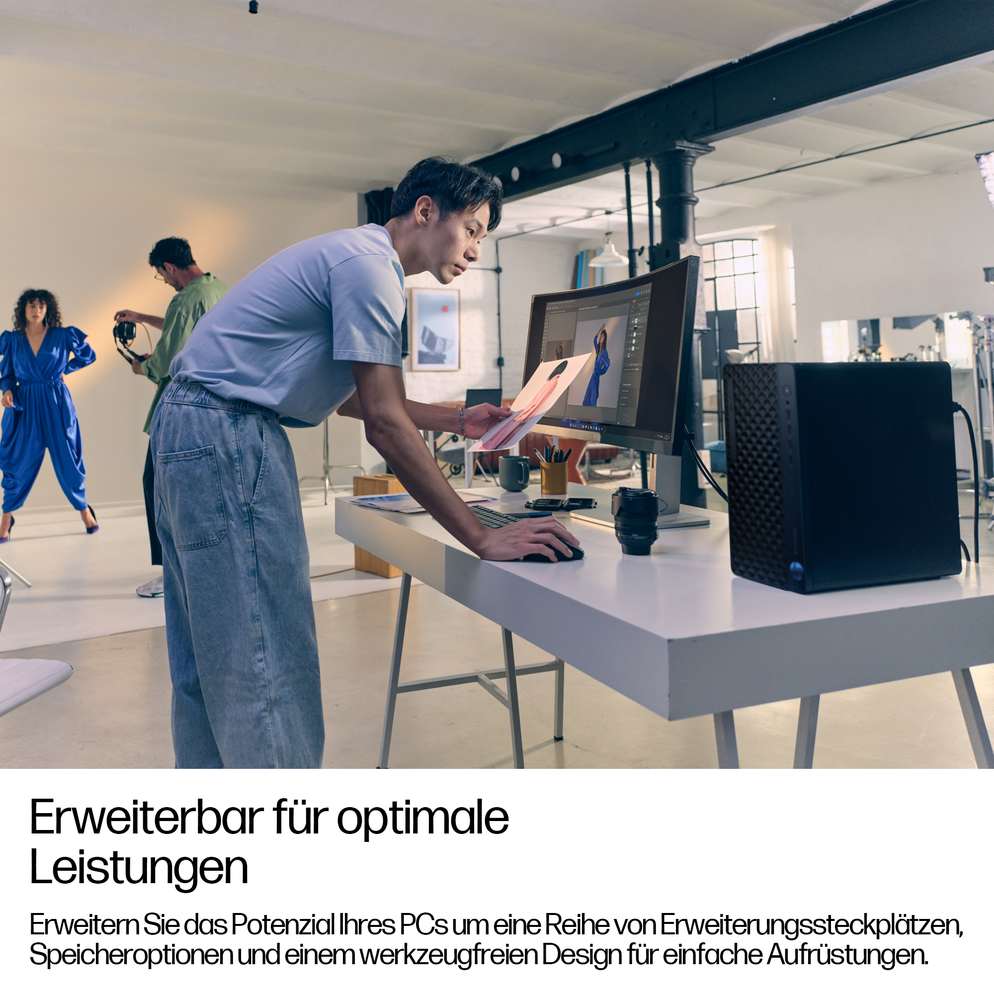Ein Mann betrachtet ein Foto auf einem Schreibtisch mit Computer und schwarzem PC. Im Hintergrund eine Frau und ein anderer Mann.