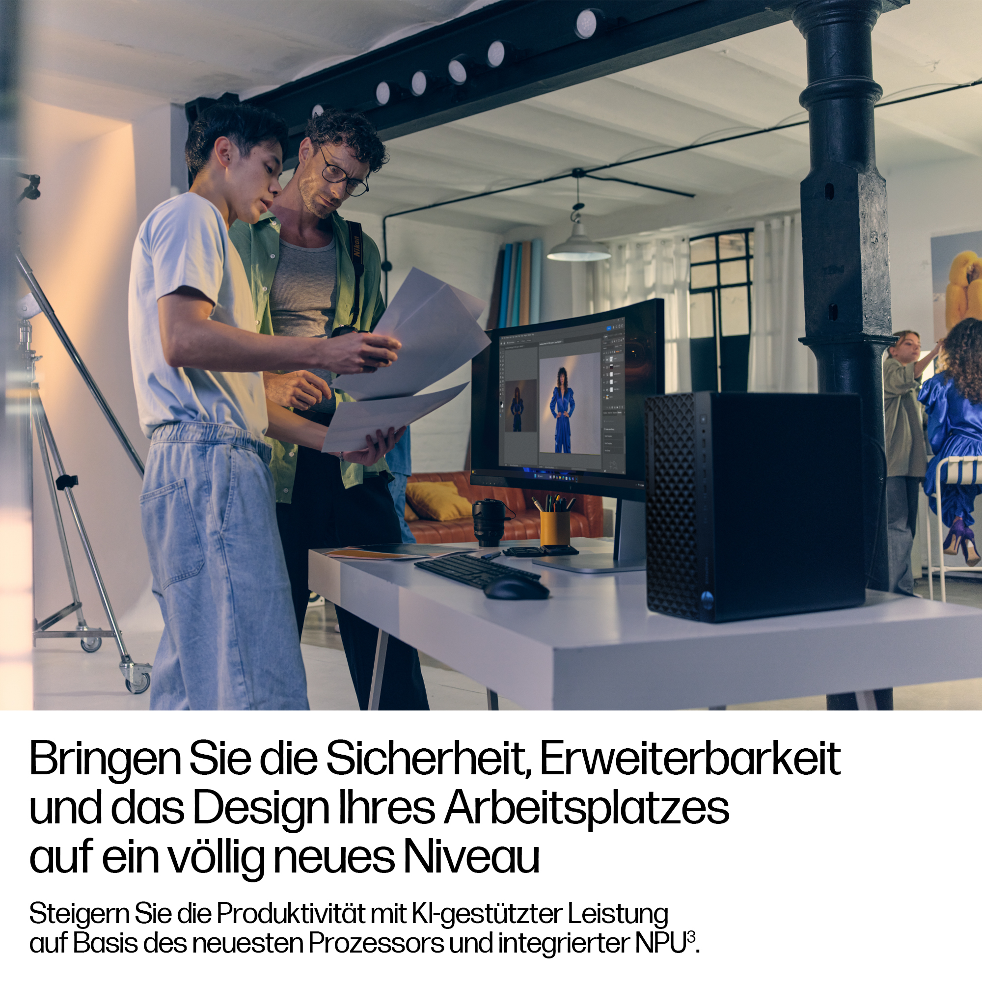 Zwei Männer untersuchen Papiere in der Nähe eines Computers mit einem Bild auf dem Bildschirm, einem Desktop und anderen Personen im Hintergrund.