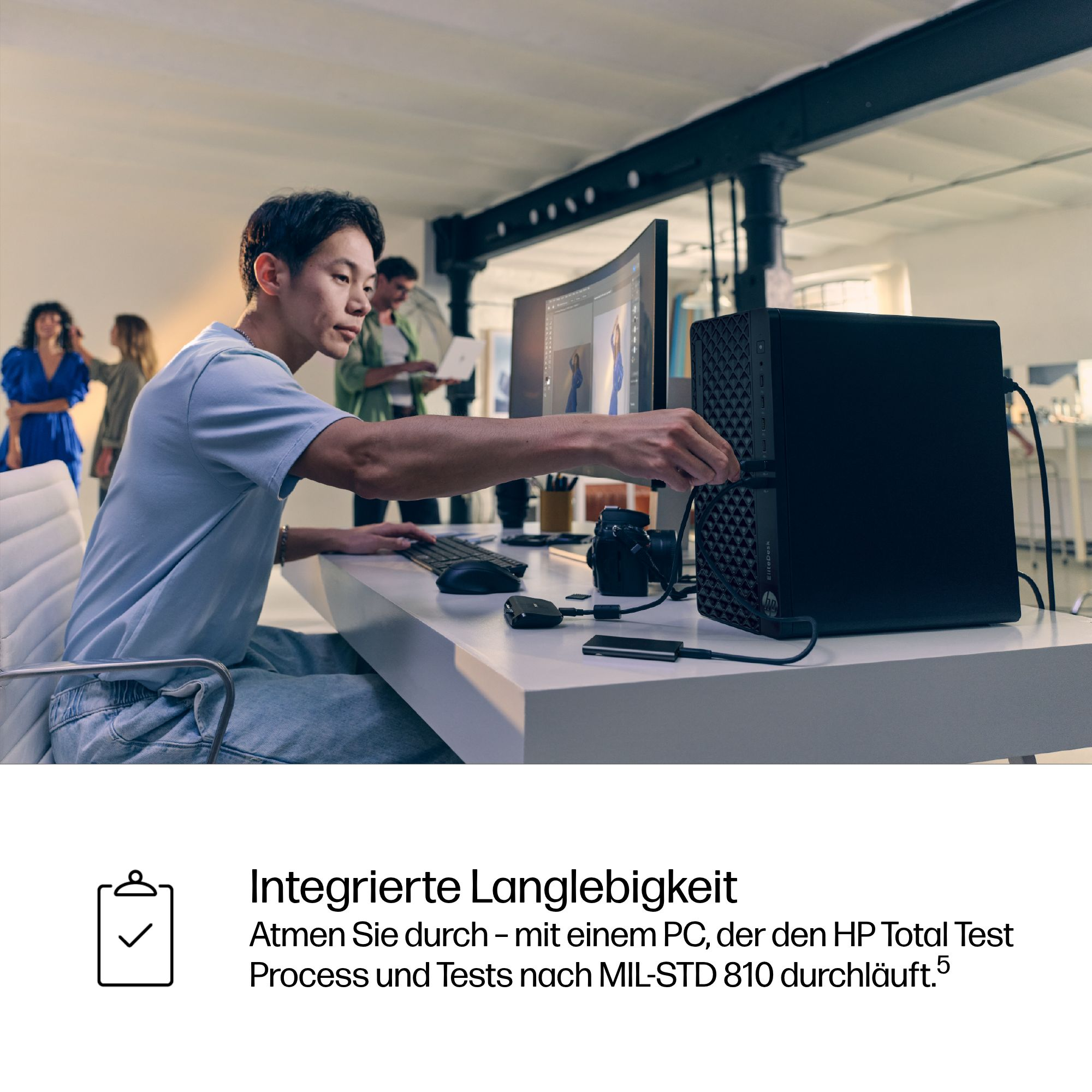Ein Mann verbindet ein Kabel mit einem schwarzen Desktop-Computer auf einem weißen Schreibtisch. Andere Leute sind im Hintergrund.
