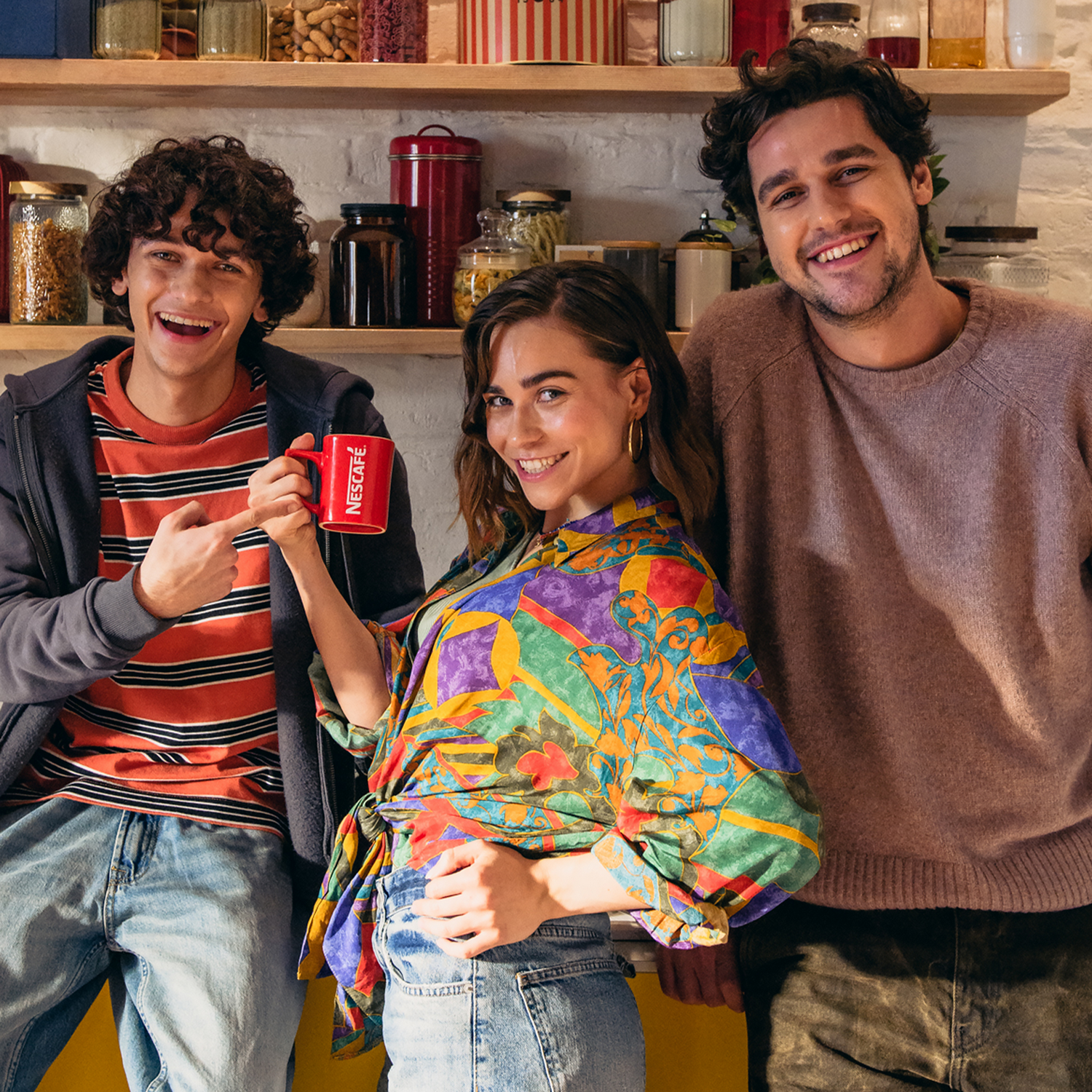 Tres personas sonriendo en una cocina. Uno sostiene una taza roja. Estantes con frascos al fondo. Camisas coloridas.
