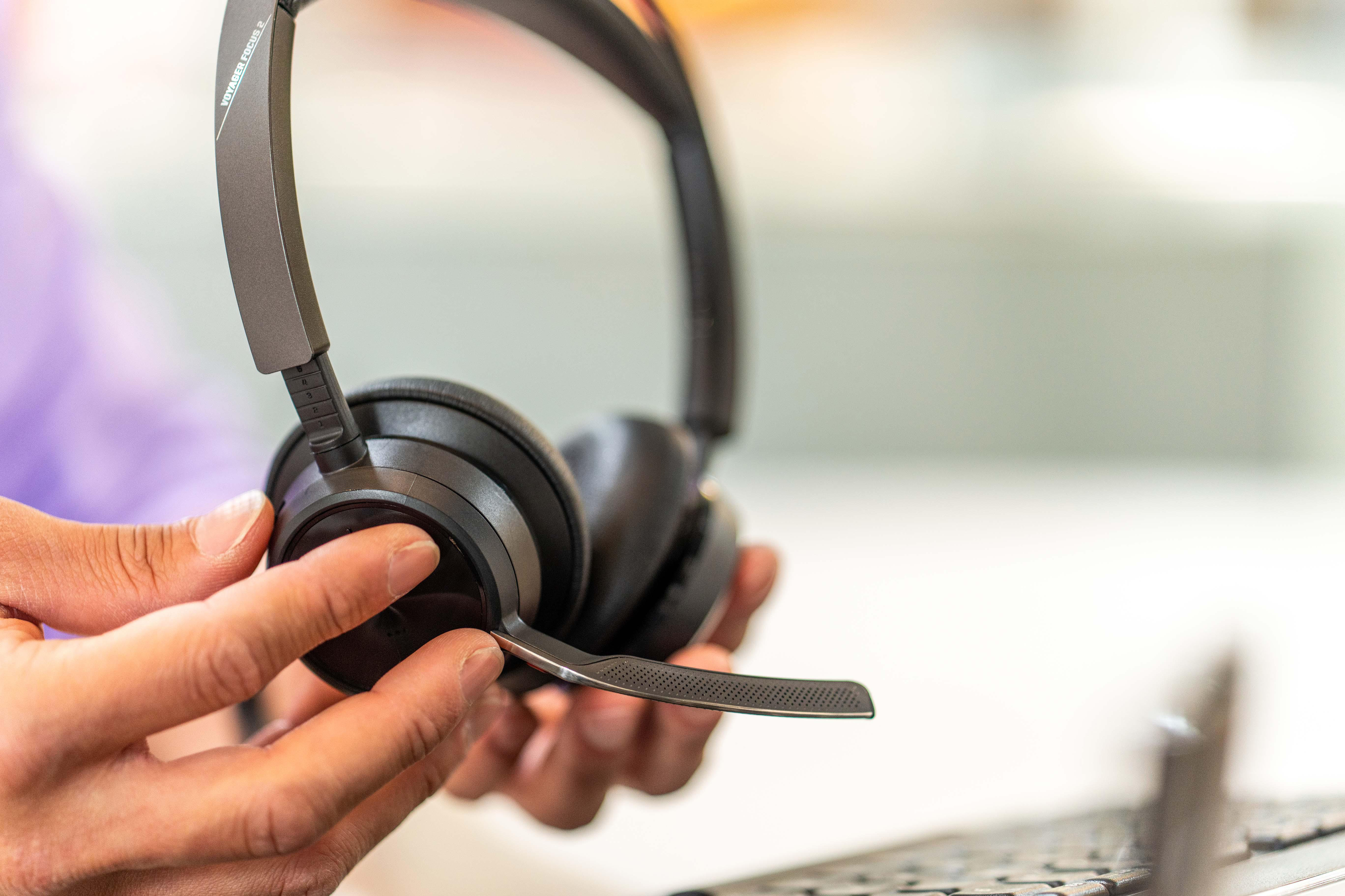 Hände halten ein schwarzes Headset mit Mikrofon. Der Hintergrund ist verschwommen und zeigt eine Tastatur und Lila.