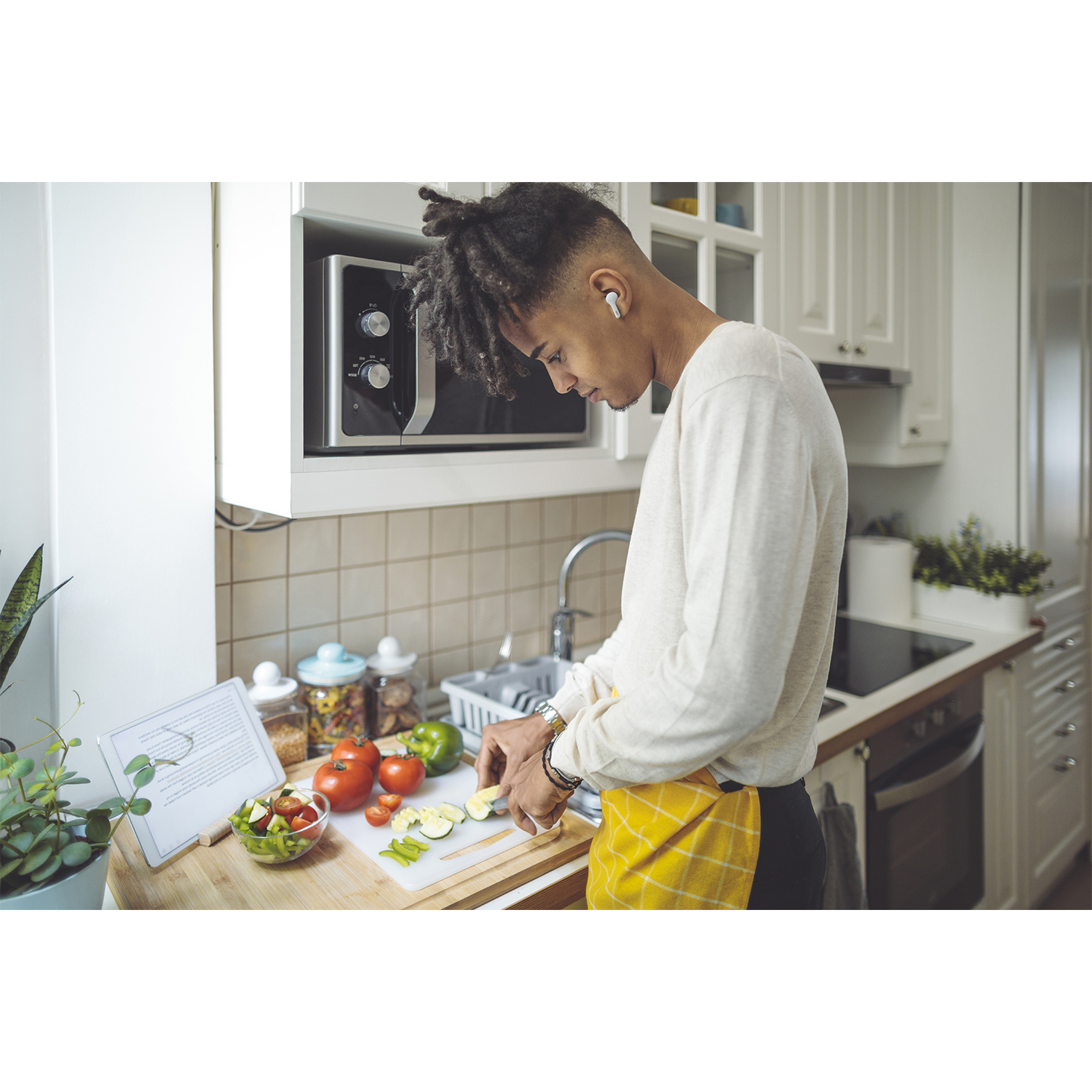 Un uomo in cucina taglia verdure. Indossa una camicia bianca, un grembiule giallo e auricolari. Ci sono pomodori, un tagliere e un microonde.