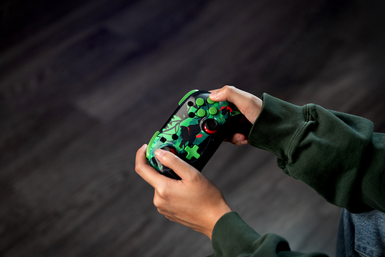 Manos sosteniendo un mando de juego con diseño verde y negro. Mangas de sudadera verde oscuro y suelo de madera en el fondo.