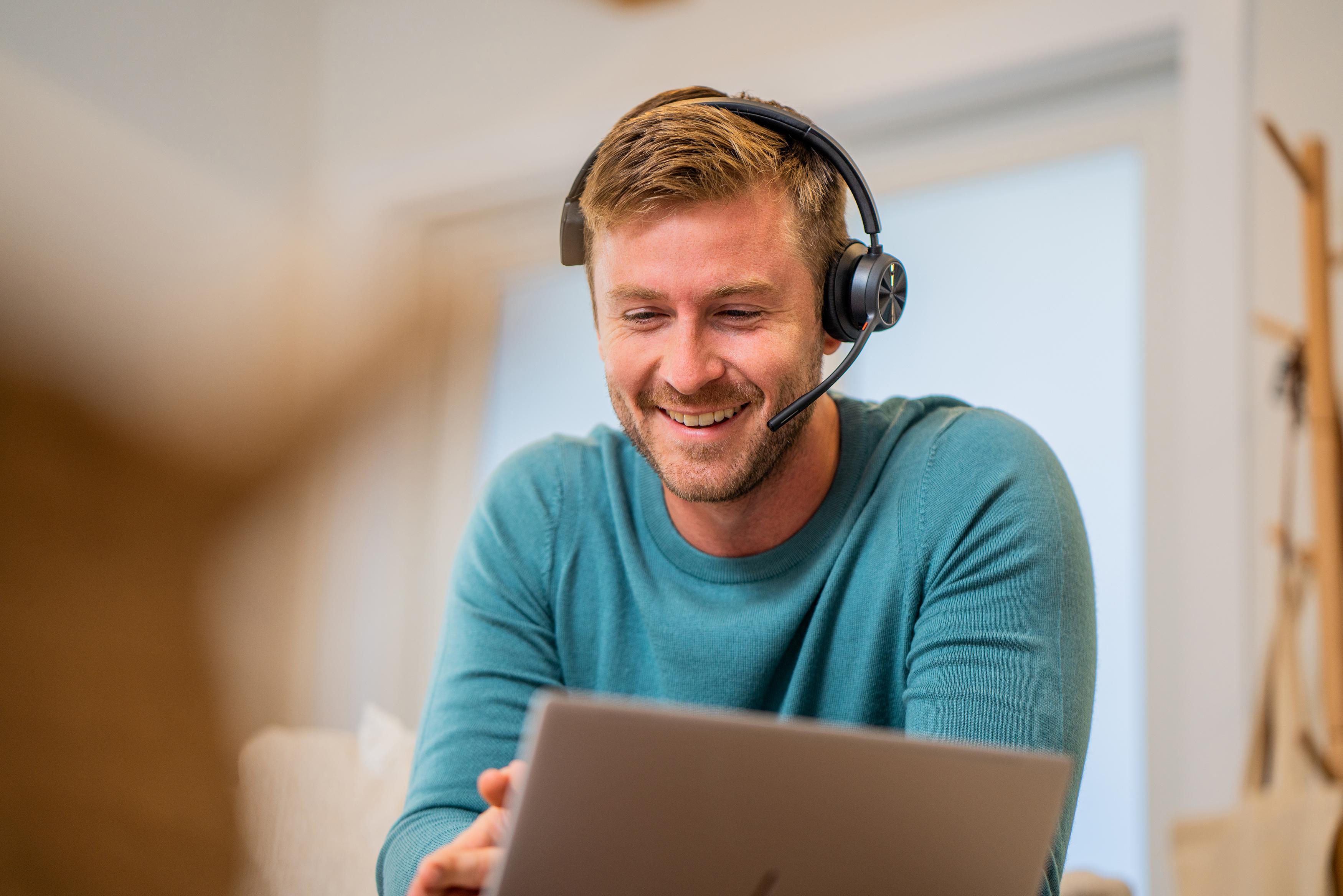 Ein Mann mit Headset lächelt einen Laptop an. Er trägt einen teal-farbenen Pullover.