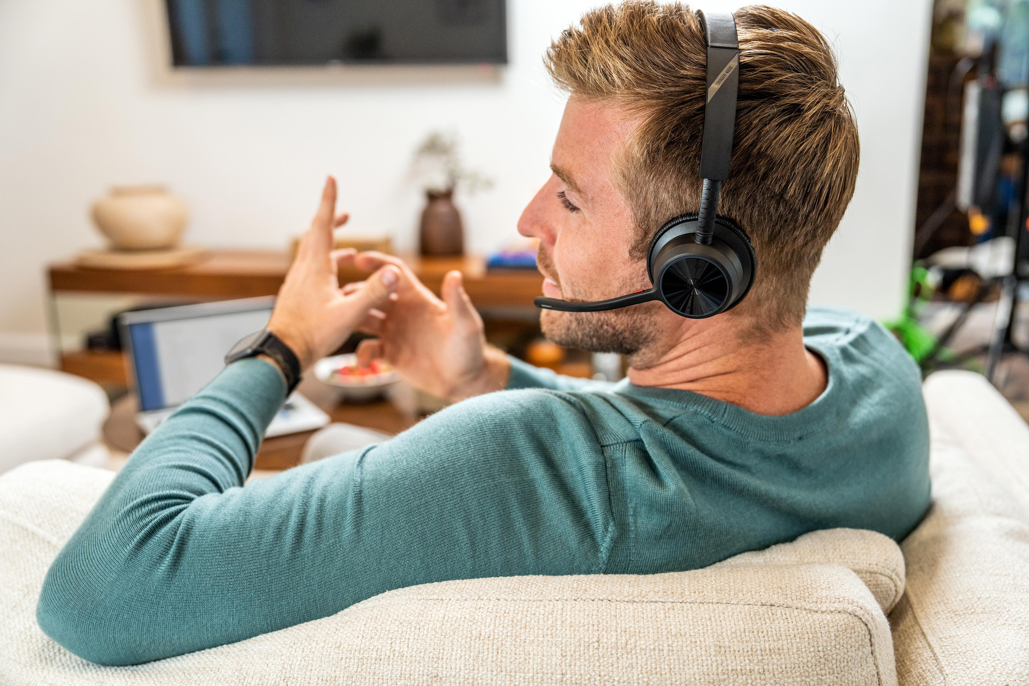 Mann mit Headset gestikuliert während eines Anrufs. Er trägt einen tealfarbenen Pullover und sitzt auf einem beigen Sofa.
