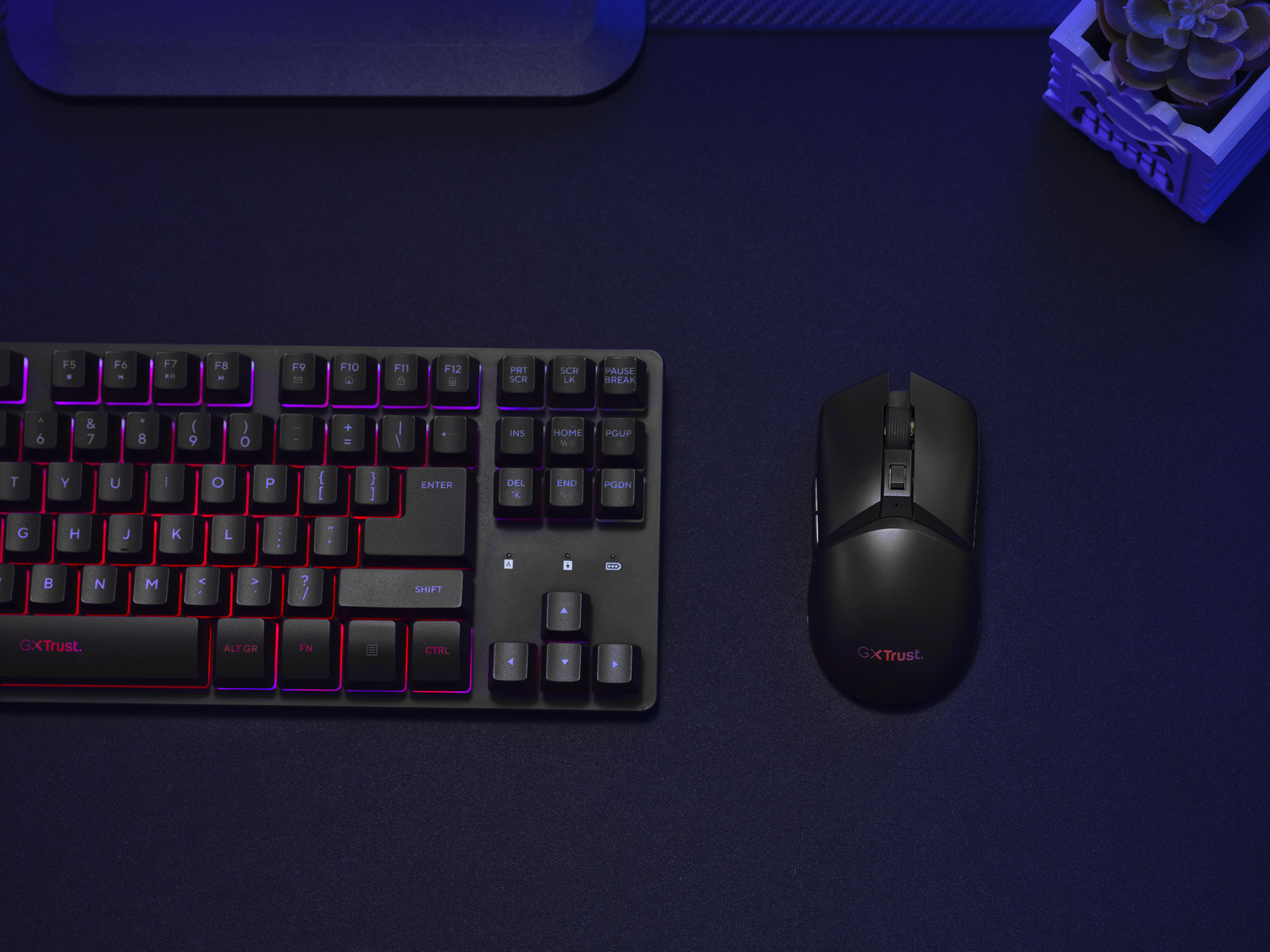 Un teclado y un ratón negros sobre una superficie oscura. El teclado tiene retroiluminación roja y morada, y el ratón el logotipo de GXTrust.
