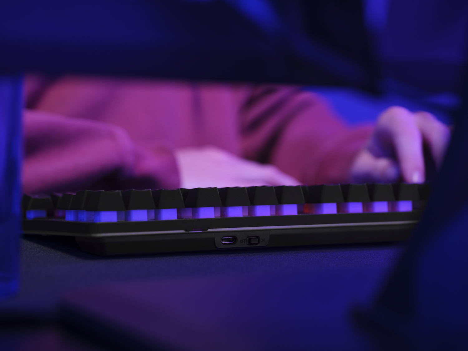 Primer plano de un teclado negro con retroiluminación púrpura. Se ven las manos y los brazos de una persona usando el teclado.