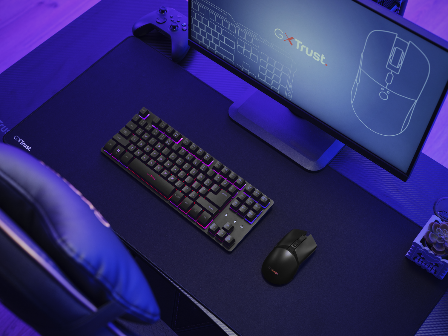 Una configuración de juego con teclado, ratón, monitor y gamepad sobre un escritorio azul oscuro. El teclado tiene iluminación colorida.