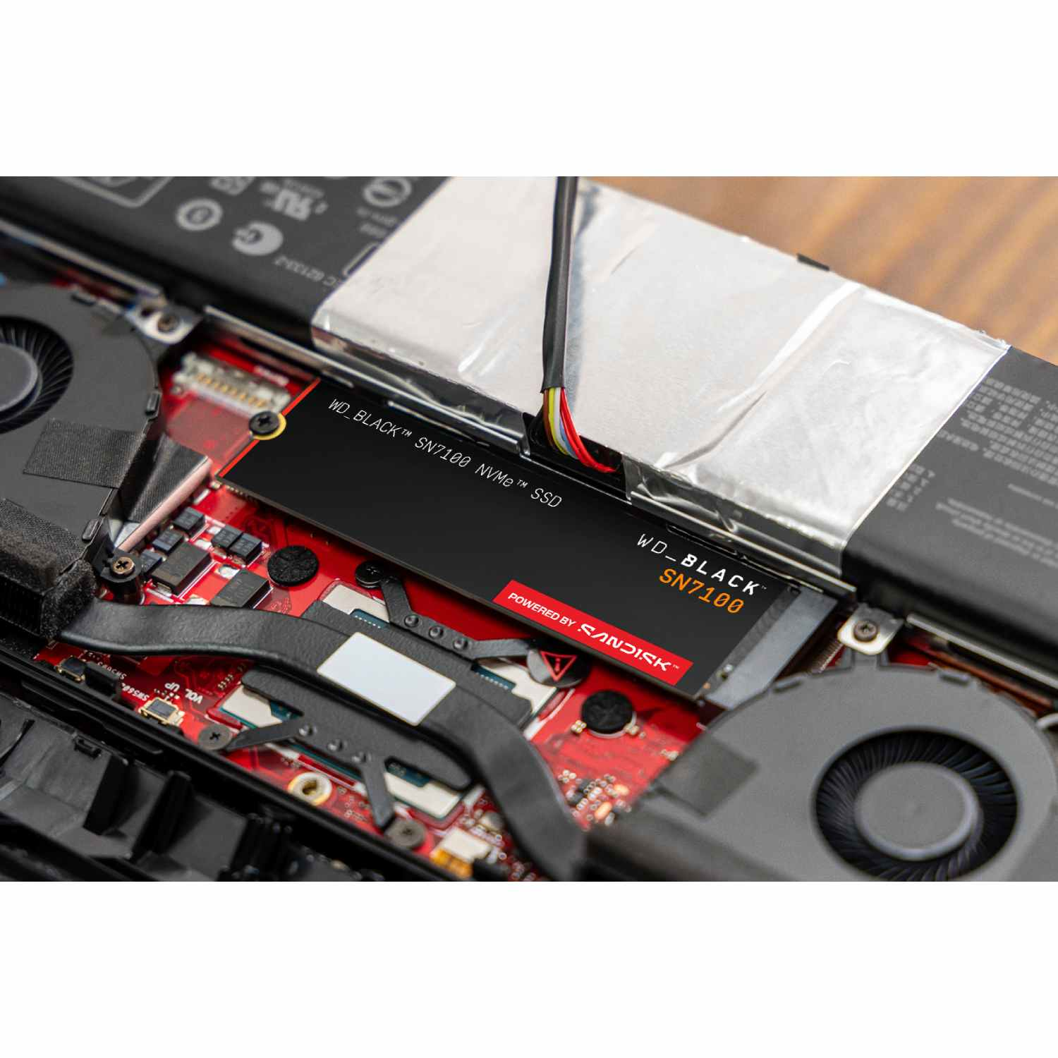 Una SSD WD_BLACK SN7100 NVMe negra está instalada en una placa de circuito rojo dentro de una laptop. Se ven cables y un ventilador.